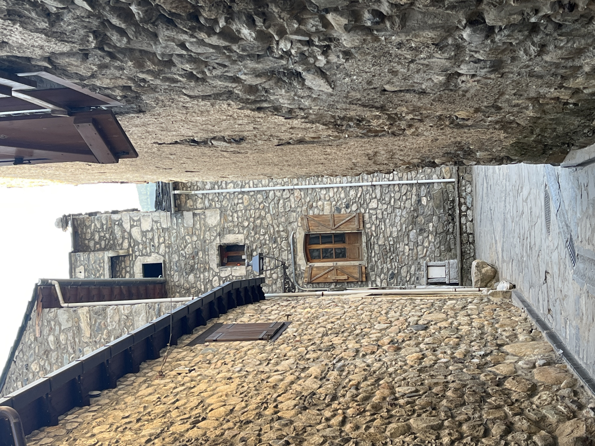 A narrow cobblestone alleyway is flanked by tall, rustic stone buildings in Benasque, Spain. The building on the left is constructed of rough-hewn, tan stones with a dark, sloped roofline and a visible gutter. A wooden window shutter is open on this building. The building on the right is closer and taller, with a facade of larger, darker, and more irregularly shaped stones, giving it a rougher texture. A wooden shutter is also visible on this building, angled outwards.
Further down the alley, a stone building with a more uniform stone construction rises. It features a single window with wooden shutters and a wrought-iron lamppost attached to its facade. Above this, another small, dark window is visible. A white pipe runs vertically along the side of this building. The alleyway itself is paved with irregular cobblestones, showing signs of wear and drainage grates. The overall scene depicts a historic and charming streetscape characteristic of old European towns.