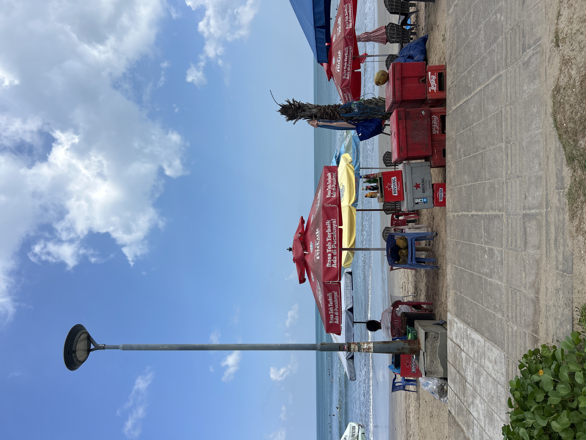 The image shows a view of Kuta Beach in Indonesia on a sunny day with a partly cloudy sky. In the foreground, there is a paved walkway with some greenery. Behind the walkway, the sandy beach is visible with several beach chairs and umbrellas set up.
Several red and yellow umbrellas are open, with signage that appears to be in Indonesian. Underneath the umbrellas, there are drink coolers, including red Coca-Cola branded coolers and grey Bintang Crystal coolers. Some bottles of drinks are also visible.
A solitary person is seated on a red chair, facing away from the camera towards the ocean. The ocean is visible in the background, with small waves and several surfers in the water. A tall lamppost stands on the left side of the image, and a dead palm tree is visible behind some of the umbrellas. The overall scene suggests a typical day at a popular beach resort.