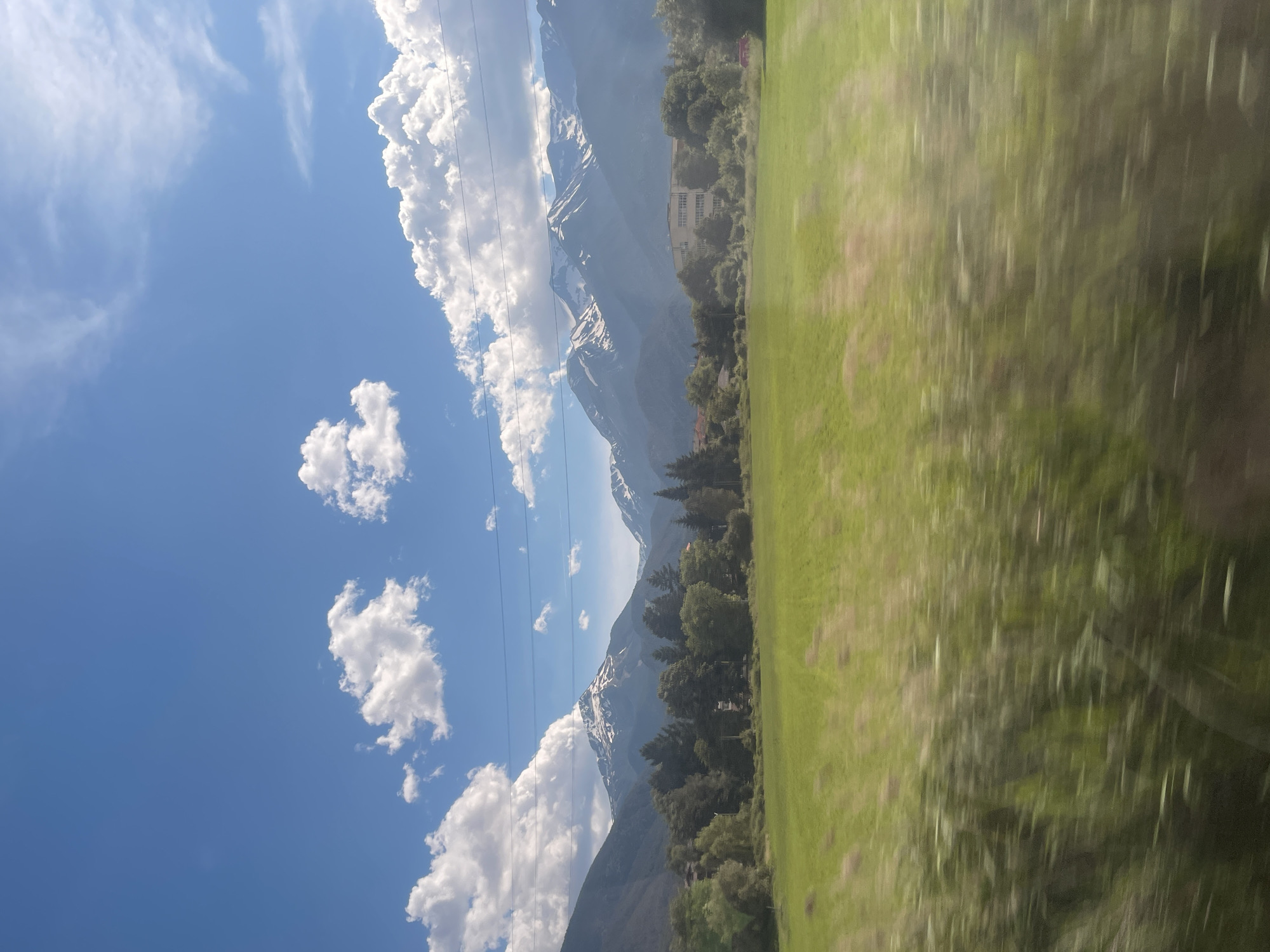 A wide shot captures a mountain landscape under a bright blue sky with scattered white clouds. The foreground is a blurred green field, suggesting motion, likely from a vehicle. Behind the field, a line of trees forms a dense border, with a few buildings partially visible among them. The background is dominated by a range of majestic mountains, their peaks capped with snow, indicating a high altitude or a cooler season. The lighting suggests it is daytime, likely midday.