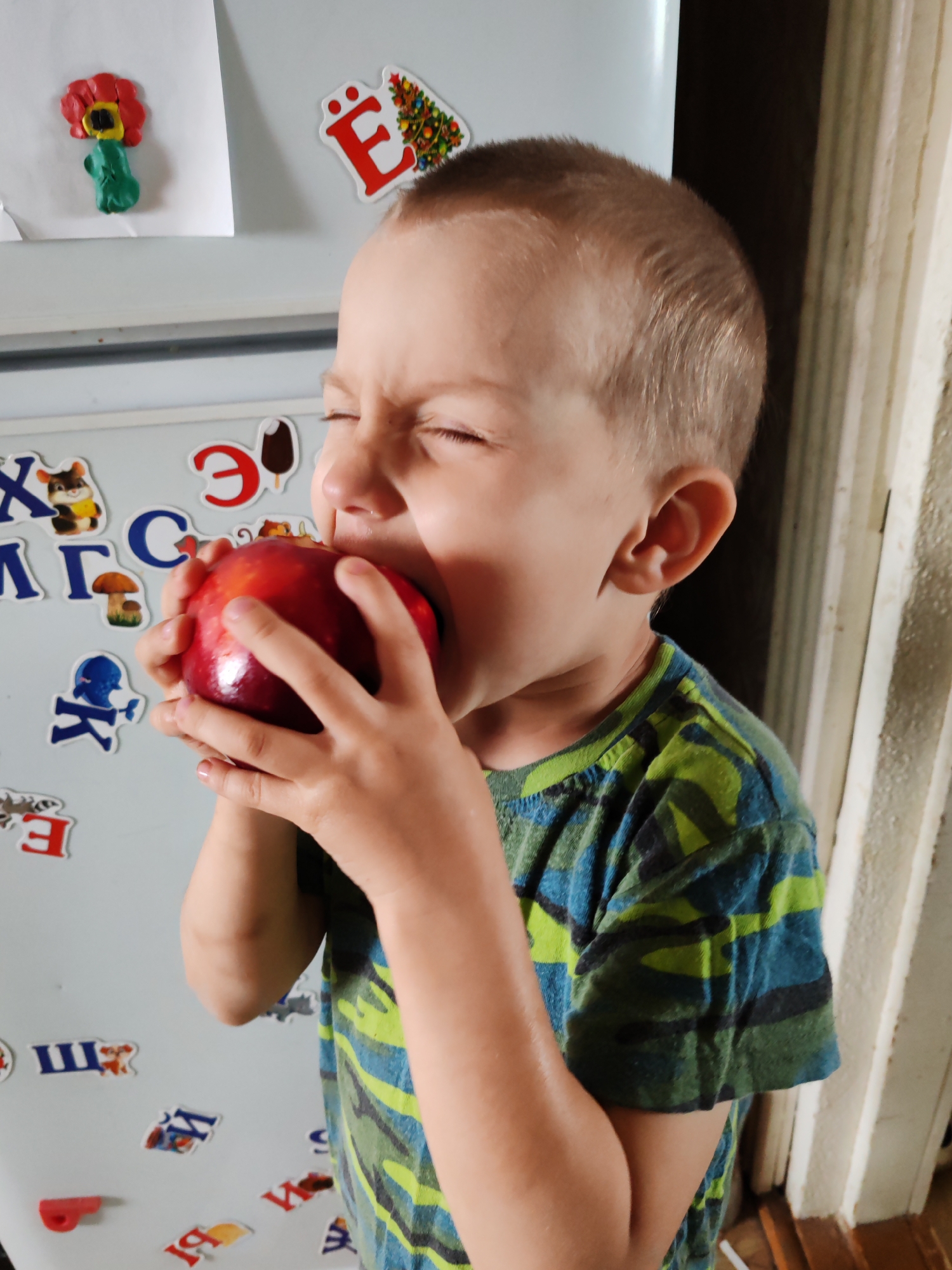 A young boy with light skin and short blond hair is eating a red apple with visible enthusiasm. He is wearing a green and blue patterned shirt. The scene appears to be indoors, possibly a kitchen, as indicated by the presence of a refrigerator in the background covered with various magnets, stickers, and a paper flower. A door frame or architectural element is visible on the right. Based on visual evidence of Russian Cyrillic letters on the refrigerator magnets, this may be in a Russian-speaking location. While the prompt requests a specific city, Чухлэм, Russia, there is no identifying data within the image to forensically confirm the image's location.