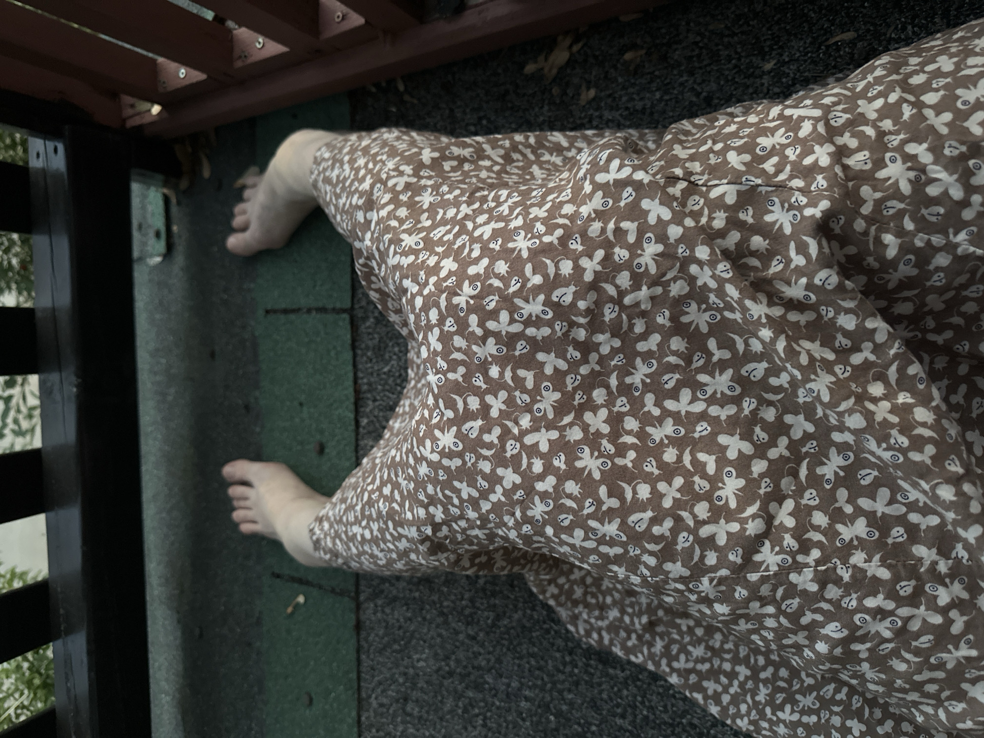 The image shows a person's bare feet and legs from a top-down perspective, wearing a brown dress with a white floral pattern. The person is sitting or lying down on a textured surface, possibly a wooden deck or patio, with some green roofing material visible beneath them. Dark wooden beams and railings are present in the background, suggesting an outdoor setting. The lighting indicates it is daytime, but potentially overcast or late in the day. The scene does not provide any specific landmarks or visual cues to determine its exact location within McMasterville, Canada.