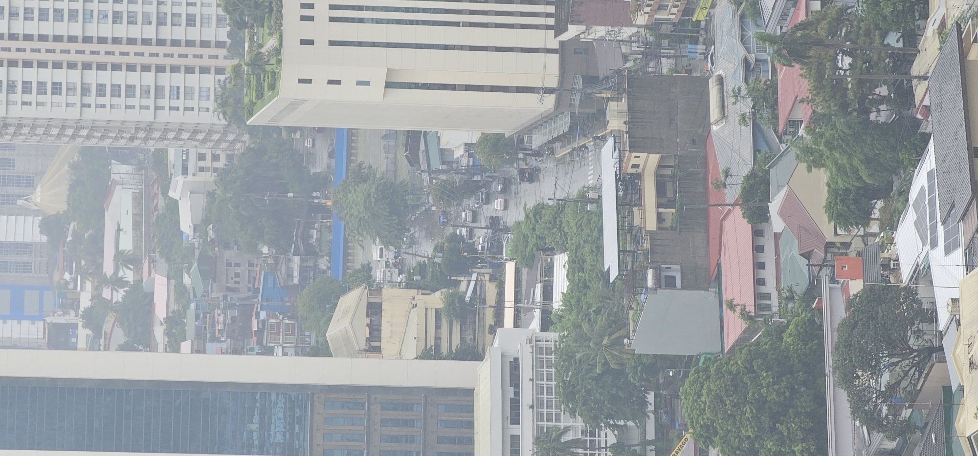 This is an aerial view of a dense urban environment in Makati City, Philippines, likely taken during a hazy or overcast day, as evidenced by the soft lighting and muted colors. The scene is dominated by a mix of modern high-rise buildings and lower-rise residential or commercial structures, interspersed with abundant greenery.
In the foreground, a prominent modern building with a glass facade and light-colored vertical accents is visible on the left. To its right, a lighter colored, boxy building with multiple windows rises, featuring a rooftop garden with palm trees. Further to the right, a tall, rectangular building with a pattern of dark windows and lighter vertical stripes dominates the mid-ground.
Below these taller structures, a network of streets and roads can be observed, with a moderate amount of vehicular traffic, including cars and possibly buses. The roads appear wet, suggesting recent rain. Several smaller buildings with various roof colors, including red, green, and light brown, are scattered throughout the scene. These buildings exhibit a range of architectural styles, from simple rectangular structures to those with more complex rooflines.
Trees are a significant element, with dense foliage creating pockets of green throughout the urban landscape. Some larger trees with visible trunks are present, as well as clusters of smaller trees and shrubs. In the background, the city stretches out, with more high-rise buildings and structures fading into the atmospheric haze. The overall impression is one of a bustling, multifaceted city with a blend of the old and the new, and a noticeable presence of nature within its built environment.