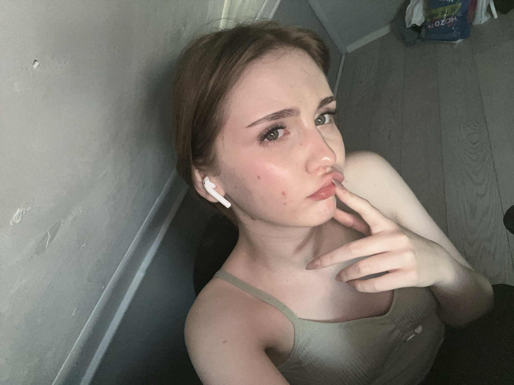 The media file is a close-up, high-angle selfie of a young woman with light skin and light brown hair. She is wearing a light green tank top and white earbuds. Her left hand is resting on her chin, and her right hand is near her mouth, with her index finger touching her lip. She has a neutral to slightly pouty expression. The background shows a gray wall with some texture and a wooden floor. In the upper right corner, there is a blurry blue bag with Russian text that appears to be "OZON." The lighting suggests it might be daytime, indoors. The overall impression is a casual, personal photograph taken in a home environment. The setting appears to be indoors, possibly a bedroom or living area, given the wall and floor textures. The woman's pose and facial expression suggest a moment of reflection or a casual pose for a selfie. There are no strong indicators of specific weather or a particular event. The presence of the "OZON" bag hints at a Russian context, consistent with the provided location information.