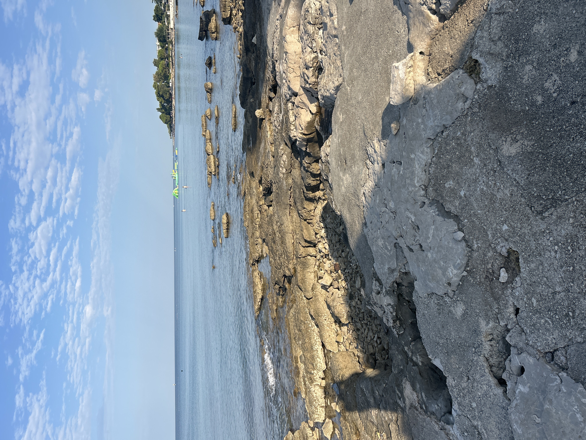 The image shows a rocky coastline in Zambratija, Croatia, with calm, blue water lapping at the shore. In the distance, a green inflatable water park is visible, along with a few people swimming or wading in the sea. Trees and buildings line the coast behind the water. The foreground is dominated by weathered, grey rocks and pebbles, with the shadow of a person cast across them, suggesting a sunny day. The sky is a pale blue with scattered white clouds.