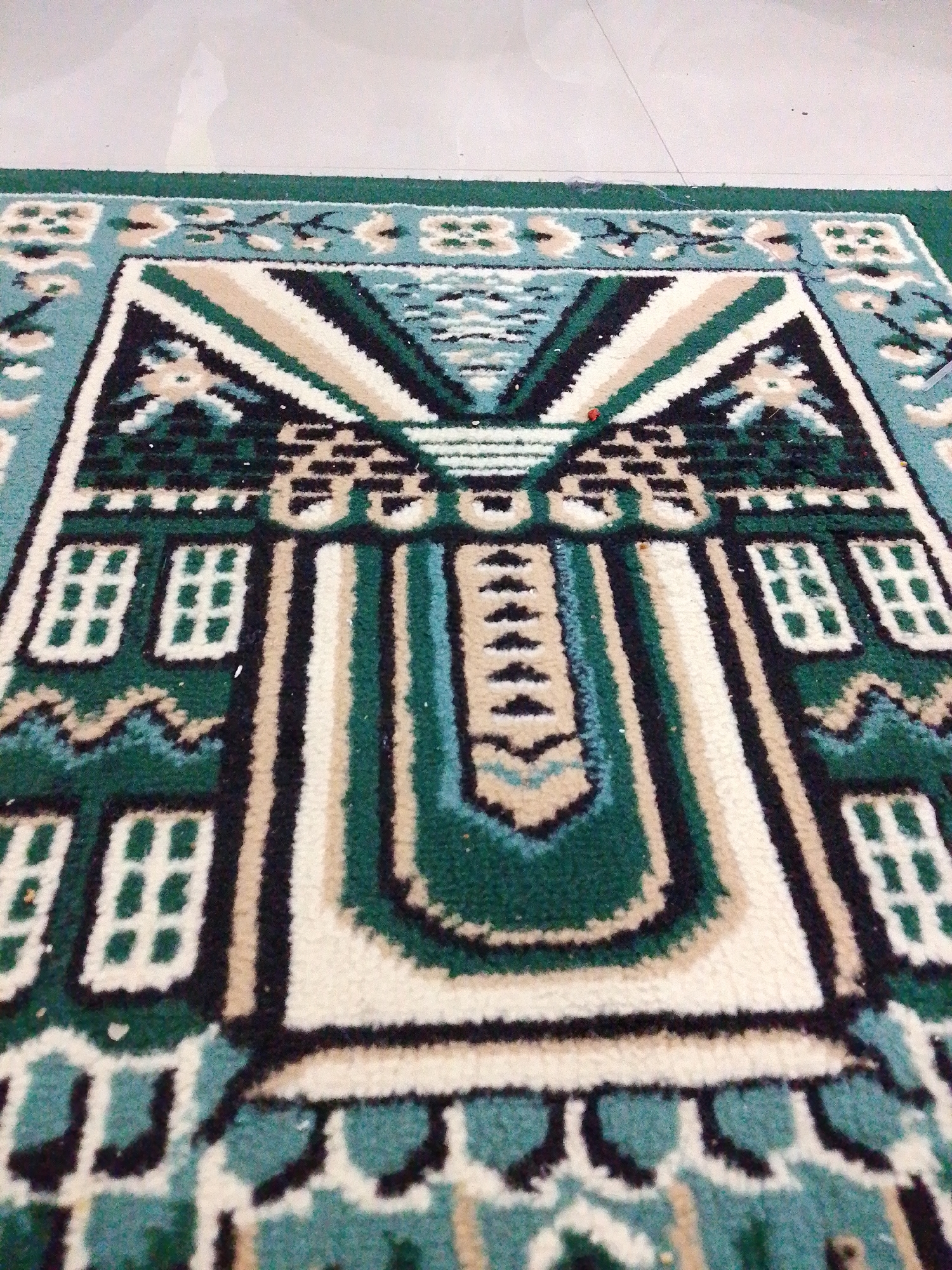 The image shows a close-up, angled view of a prayer rug placed on a tiled floor. The rug has a detailed geometric pattern in shades of green, blue, cream, and black. The pattern includes what appear to be stylized architectural elements, possibly representing a mihrab or a mosque facade, with repeating square motifs resembling windows on either side. The rug's edges are bordered in green. The floor tiles are light-colored and appear to be smooth and reflective. The scene is static, with no people or actions depicted. The overall impression is one of a prayer space within a building. The location is the city of Lawatan, Indonesia.