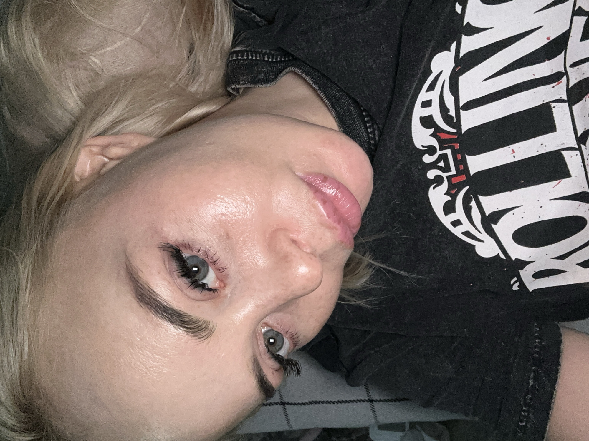 A close-up, high-angle shot captures a person with light skin, blonde hair, and blue-grey eyes, lying down and looking directly at the camera. The subject appears to be a woman, with clear skin that has a slight sheen. Her eyebrows are well-defined, and she wears prominent dark mascara or false eyelashes. Her lips are full and coated with a glossy, light pink or nude lip product.
She is wearing a dark, possibly charcoal or black, t-shirt with a bold white graphic design. Partially visible on the shirt are large white letters, with what appears to be part of the word "ROLLING" or a similar word, including visible parts of 'R', 'OLLIN', and 'G'. The design incorporates decorative white scrollwork and subtle red accents or distress marks. Her blonde hair frames the left side of the frame, and a glimpse of an ear is visible amidst the hair. In the lower left corner, a gray and white plaid or checkered fabric, possibly a blanket, is visible, suggesting an indoor setting. The lighting is soft and appears to be artificial. No specific activity is depicted other than posing for the camera with a neutral expression.
