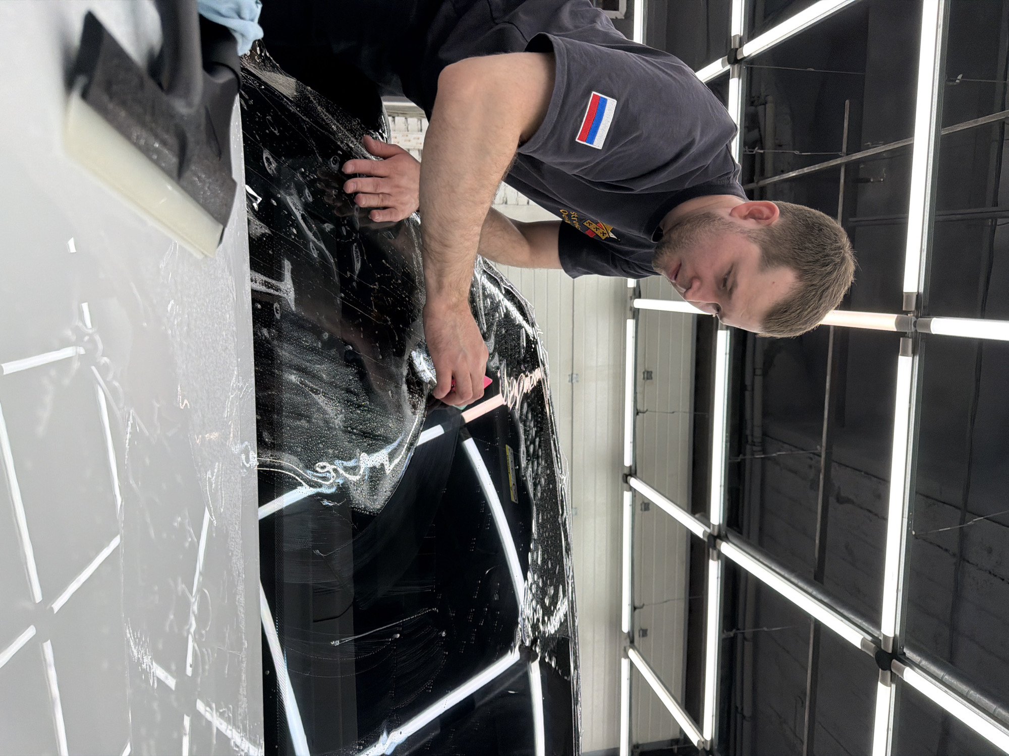 A man is applying a transparent film to the hood of a black car in what appears to be a garage or automotive detailing shop. The man is wearing a gray t-shirt with a patch resembling the Russian flag on his left sleeve. He is bent over the car, using his hands and a pink tool to smooth the film onto the surface. The hood of the car is covered in a soapy solution. To the left of the car is a white surface, possibly another part of the car, and a black squeegee is visible. The shop has rows of bright fluorescent lights arranged in a grid pattern on the ceiling. The location may be in Krasnoyarsk, Russia, inferred from the flag patch.