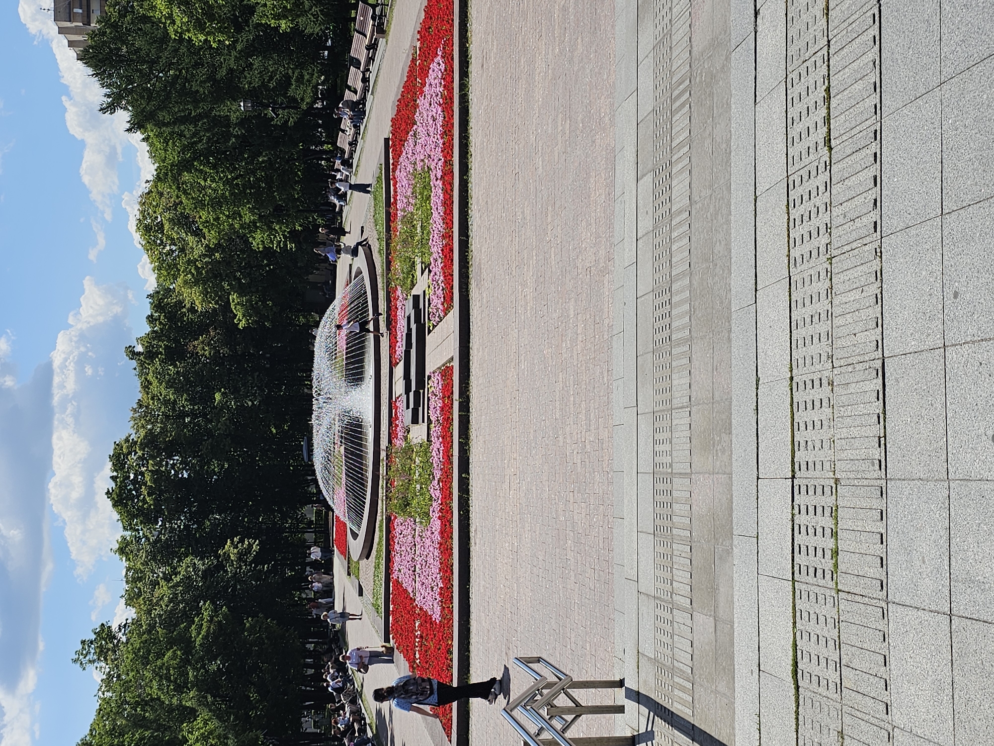 The image depicts a sunny day in a park in Moscow, Russia, likely in the vicinity of a prominent landmark, given the well-maintained and decorated setting. In the foreground, there is a paved walkway with tactile paving indicating a change in elevation, suggesting it's a public space designed for accessibility. Metal railings are visible on the left, leading down.
The midground features a large, circular fountain spraying water, with pink and red flowers arranged in geometric patterns around its base. Beyond the flowerbeds, a paved area with benches is visible, where several people are seated, enjoying the day. A cluster of trees forms a backdrop to the scene, with lush green foliage. In the distance, a multi-story building can be seen, indicating an urban environment.
Several individuals are present in the park, some walking, others sitting on benches. One person, a woman with a backpack, is walking towards the fountain. The overall atmosphere is peaceful and recreational. The lighting suggests it is daytime, with a clear sky and scattered clouds.