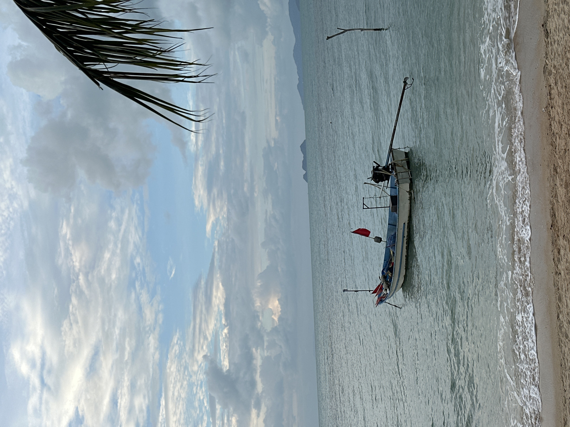 An overcast day at a beach in Koh Samui, Thailand, features a single, small motorboat anchored in calm, turquoise water. The boat, white with a blue stripe, has a red flag and is equipped with a long, dark outboard motor. Gentle waves lap against a sandy shore in the foreground, and a palm frond hangs into the frame from the upper right corner. In the distance, hazy islands and mountains form the horizon under a sky filled with soft, gray clouds. A single wooden post stands in the water to the right of the boat. There are no people or animals visible in the image.