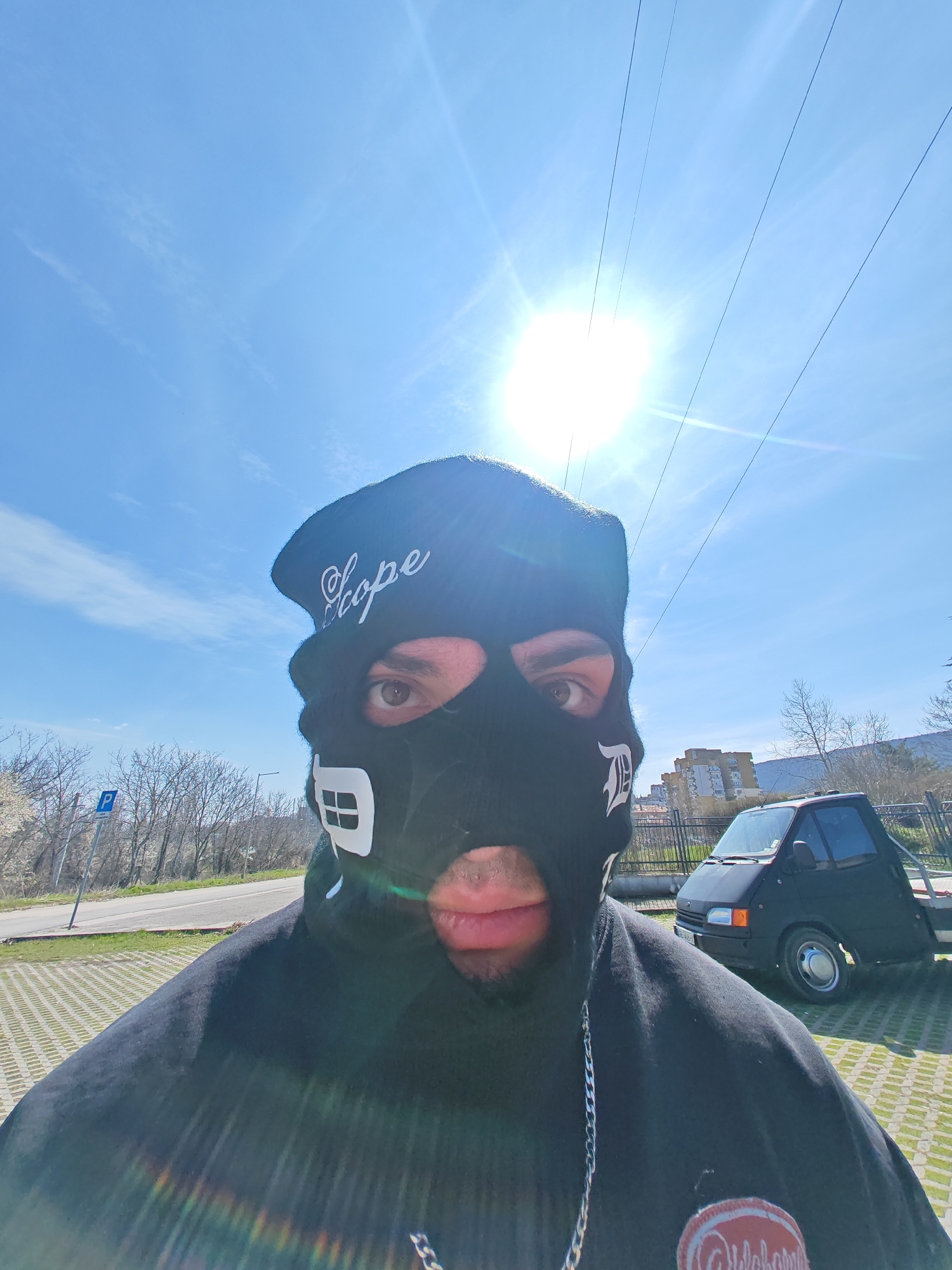 A close-up, front-facing photograph depicts an individual wearing a black balaclava and a dark hoodie on a bright, sunny day in Shumen, Bulgaria. The balaclava features white text "Scope" on the forehead, a white house-like graphic on the left cheek, and a white stylized "D" on the right cheek. Only the individual's brown eyes and lips are visible through the mask. A silver-colored chain is partially visible around the neck.
The background shows an outdoor environment. To the left, a paved parking area with a grid pattern transitions to a narrow asphalt road. A blue rectangular sign with a white 'P' (parking symbol) is visible roadside. Beyond the road, deciduous trees with bare branches line the area. To the right, a compact, dark-colored utility vehicle or mini-truck with a flatbed is parked on a similar paved surface. Behind the vehicle, a dark metal fence runs along the perimeter, followed by more bare trees and a multi-story residential building in the distance. The sky is clear blue with a prominent, bright sunburst in the upper right quadrant, accompanied by visible power lines stretching diagonally across the frame.