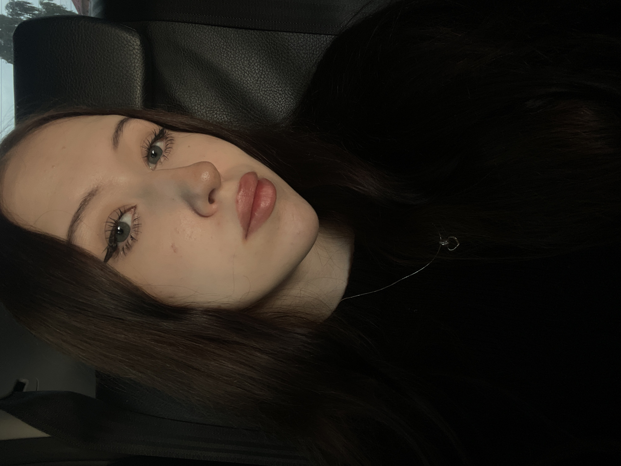 A young woman with long, dark brown hair is pictured from the chest up, looking directly at the camera. She is wearing a black top and a thin silver necklace with a small heart-shaped pendant. Her makeup includes winged eyeliner and lip gloss. The background appears to be the interior of a car, with a black leather seat visible behind her, and a glimpse of greenery outside a window. The lighting is dim, suggesting it is either nighttime or an overcast day. The image was taken in Gavrilovka, Russia.