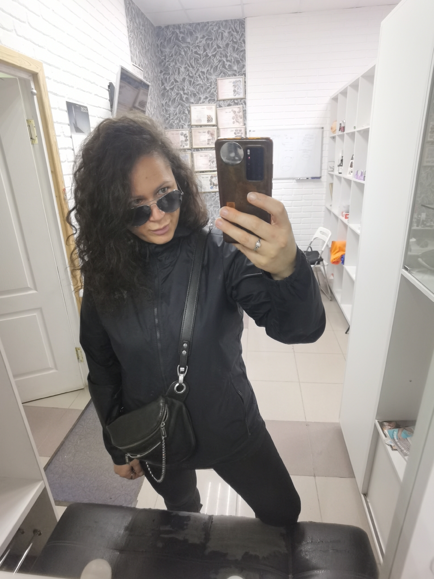 The image shows a woman taking a selfie in a room with white brick walls and shelves filled with items. She is wearing sunglasses, a black jacket, black pants, and a black crossbody bag with a chain strap. She is also wearing a ring on her left hand. In the background, there are framed certificates on the wall and a whiteboard. The room appears to be a retail space or office, possibly a salon or a shop. The lighting suggests it is daytime. The woman's expression is neutral, and she is looking at the camera. There is no visible text in the image. The location is Chaikovsky, Russia.