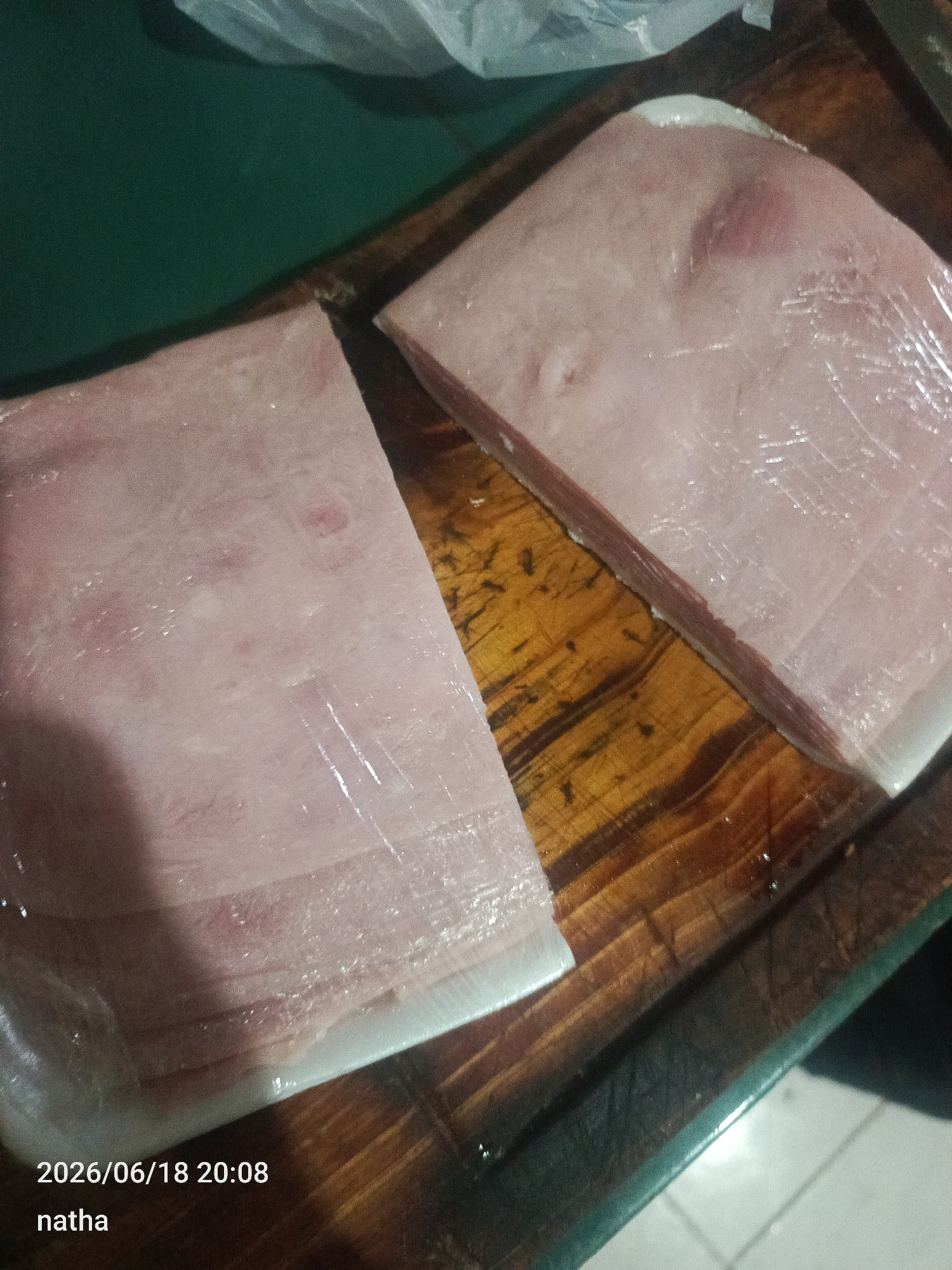 The image shows two blocks of raw ham, wrapped in plastic, resting on a wooden cutting board. The ham is light pink in color, with visible streaks of fat. The cutting board has a dark, weathered appearance, with visible grain lines. A green surface is visible in the upper left corner of the image, and a white plastic bag is partially visible behind the ham. The lower left corner of the image contains a timestamp, "2026/06/18 20:08", and the text "natha". The scene is indoors, likely a kitchen, but no specific location within El Chaparral, Venezuela can be determined from the visual information provided.