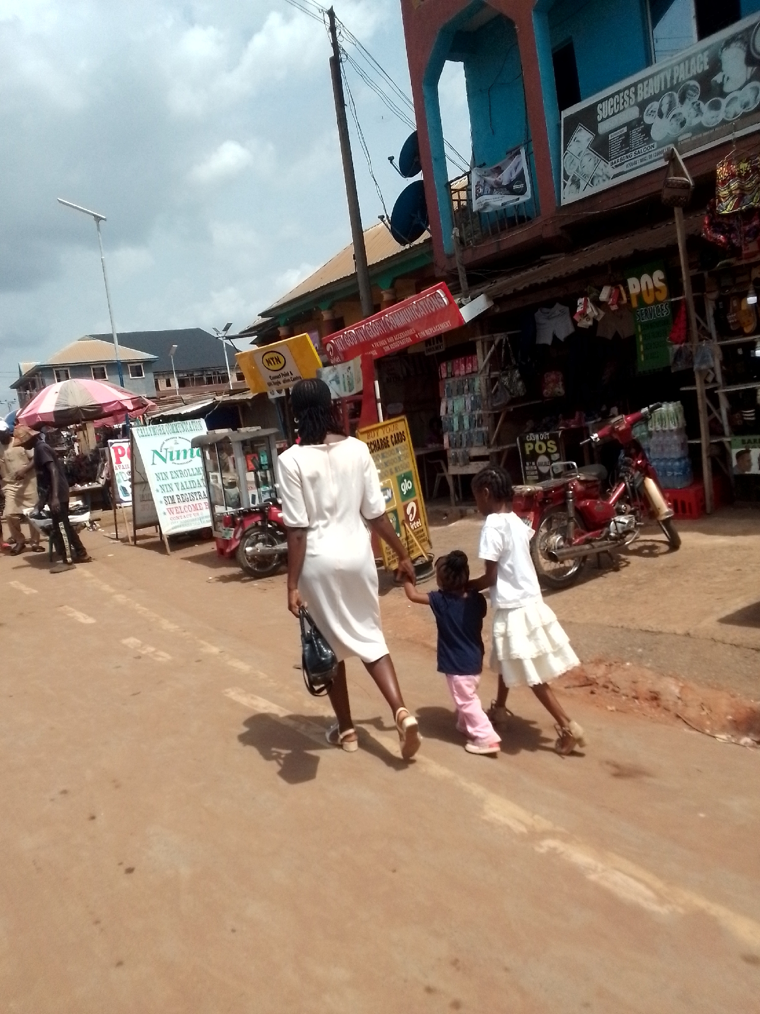 In a street scene in Orlu, Nigeria, a woman in a white dress walks away from the viewer, holding hands with two young girls. One girl wears a white shirt and skirt, while the other wears a blue top and pink pants. They are walking on a paved road.
Behind them, the street is lined with open-front shops and stalls. Signs advertise services like "NIN Enrollment," "SIM Registration," and "POS Services." Brands such as MTN, Glo, and Airtel are also visible.
A red motorcycle is parked near one of the shops. The building "Success Beauty Palace" is visible on one of the upper levels. A cloudy sky is overhead.