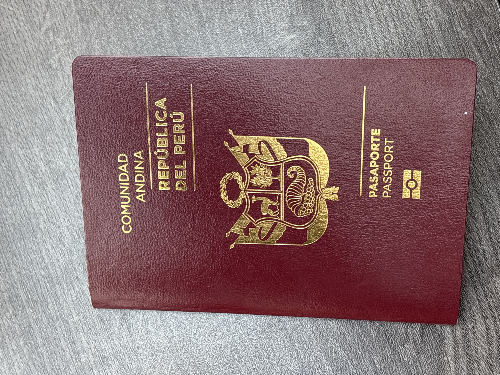 The image shows a Peruvian passport lying on a gray surface. The passport cover is a dark red color and has gold lettering and emblems. The words "COMUNIDAD ANDINA" and "REPÚBLICA DEL PERÚ" are printed vertically on the left side. In the center is the coat of arms of Peru, and to the right are the words "PASAPORTE" and "PASSPORT" printed vertically. The image does not contain any information about Puebla, Mexico.
