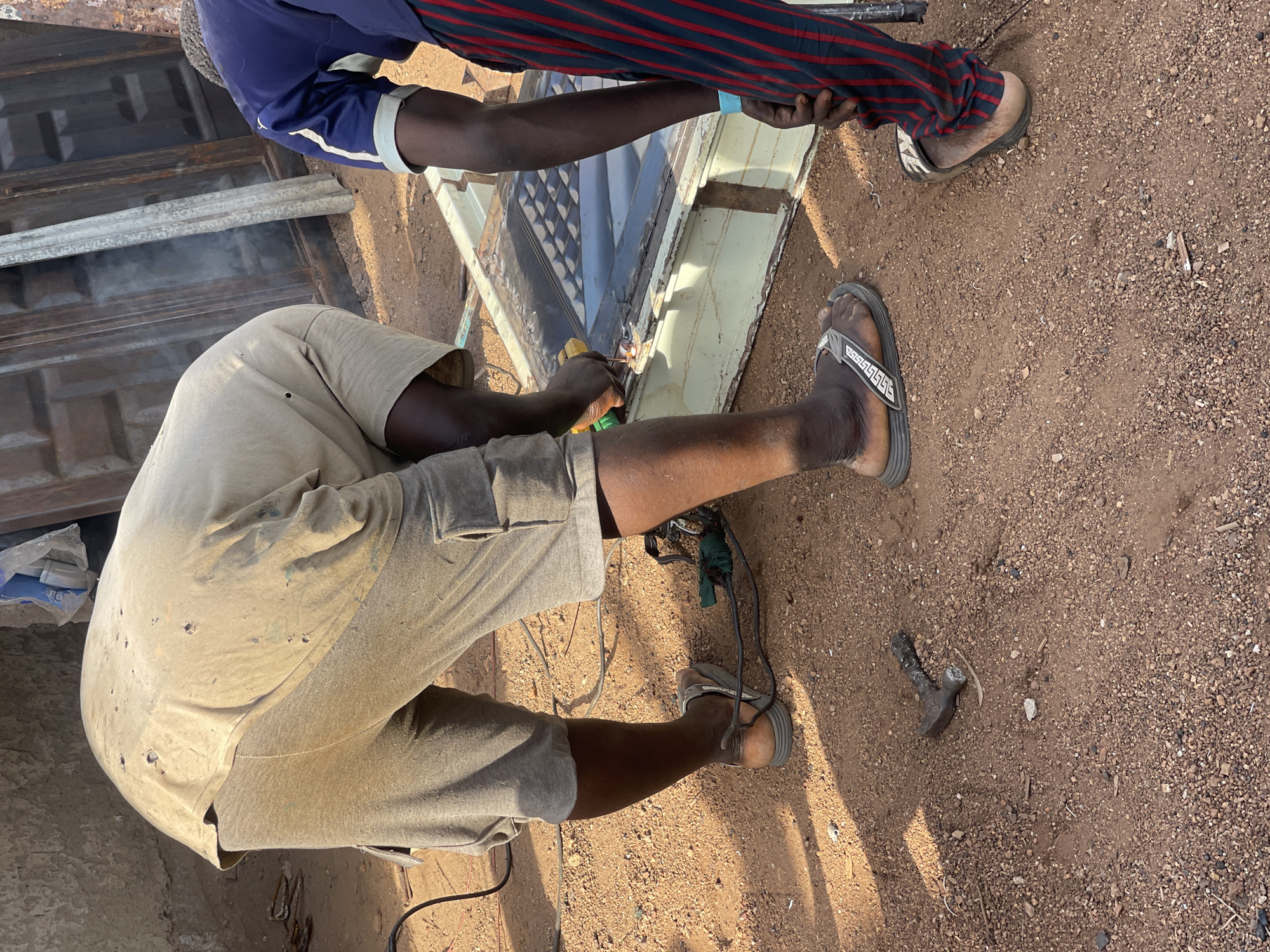 In Akwanga, Nigeria, two men are welding a metal door. One man, wearing a light-colored shirt and shorts, is welding while kneeling on the ground. The second man, wearing a blue shirt with striped sleeves, is holding the metal door in place. The ground is dirt and gravel. Tools, including a hammer, are visible near the welder.