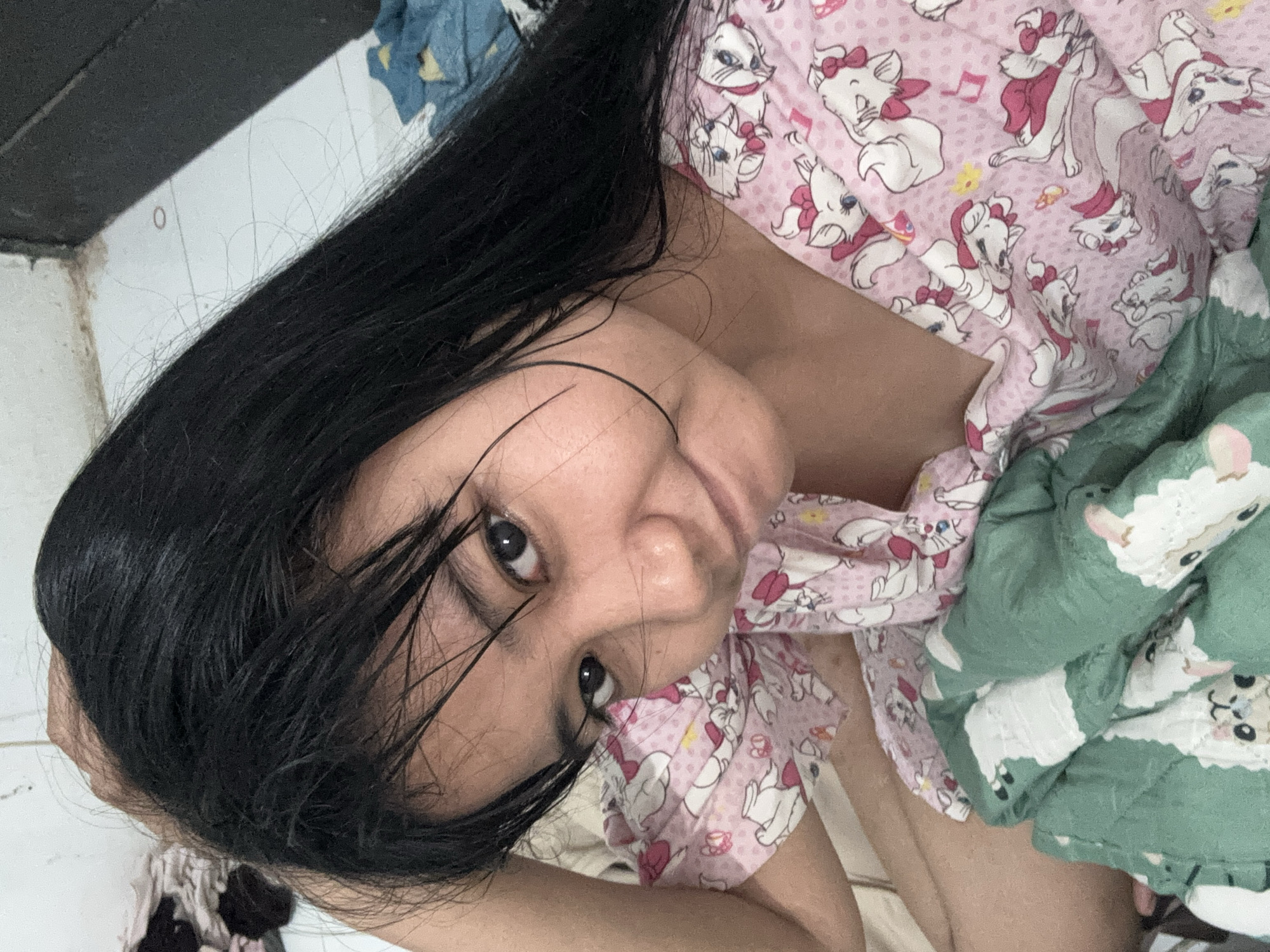 An Indonesian woman with long, dark hair lies in bed, looking up at the camera. Her hair is partially covering her face. She is wearing a pink pajama top with white cat patterns. A green blanket with white lamb patterns is draped over her. The room appears to be a bedroom. The image was taken in North Jakarta, Indonesia.
