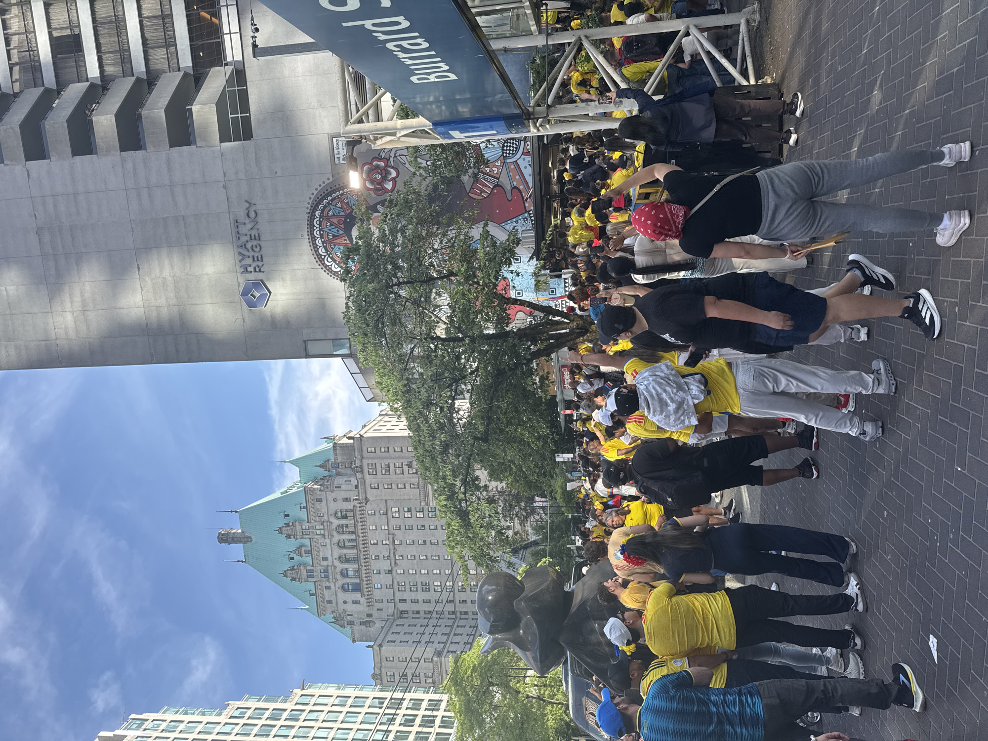 A crowd of people, many wearing yellow shirts, are gathered on a city street. In the background, there is a large, old-fashioned building with a green roof and a modern building with "HYATT REGENCY" signage. A mural is visible on the side of the Hyatt Regency building. A street sign reading "Burrard St" is also present. The people appear to be engaged in a gathering or event, with some holding up their phones and others with their arms raised. The street is paved with brickwork. The sky is blue with some clouds.