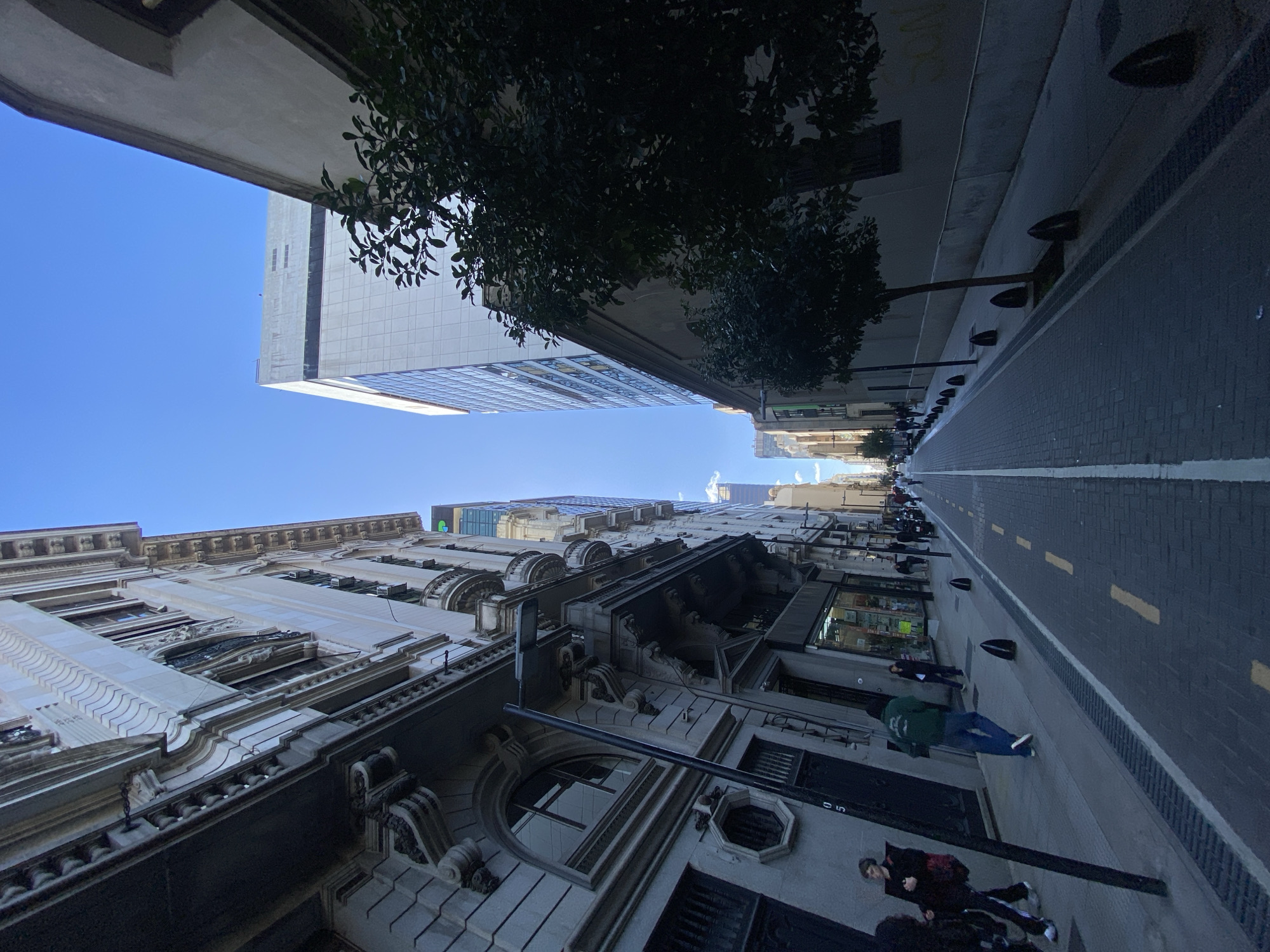 This image captures a vibrant urban street scene in Buenos Aires, Argentina, seen from a low angle looking upwards. The perspective emphasizes the towering architecture and the clear blue sky above.
On the left side of the street, a magnificent multi-story building dominates the view, showcasing ornate classical architecture. It features intricate stone carvings, decorative cornices, several balconies, and arched windows, suggesting a building of historical significance. The facade is a light, perhaps cream-colored, stone.
To the right, a mix of architectural styles is visible. A modern, glass-fronted skyscraper, reflecting the sky, stands tall, contrasting sharply with the older buildings. Patches of lush green foliage from trees line the sidewalk, casting shadows and adding a natural element to the concrete jungle.
The street itself is paved with dark cobblestones or bricks, featuring a crisp white line on one side and broken yellow dashed lines in the middle, indicating a designated traffic lane. A narrow sidewalk runs alongside, equipped with dark, rounded bollards or planters.
Several people are engaged in everyday activities on the sidewalk. In the foreground, a person in dark attire stands, while further down, another individual wearing a green hoodie and blue jeans walks away from the viewer. Other distant figures suggest a moderate level of pedestrian traffic.
The scene takes place on a bright, clear day, indicated by the brilliant blue sky and the distinct shadows cast by the buildings and trees. The overall impression is one of a bustling city combining historical grandeur with modern development. A faint green logo, possibly "Go", is visible on the modern skyscraper. The number "05" is visible on a lower facade.