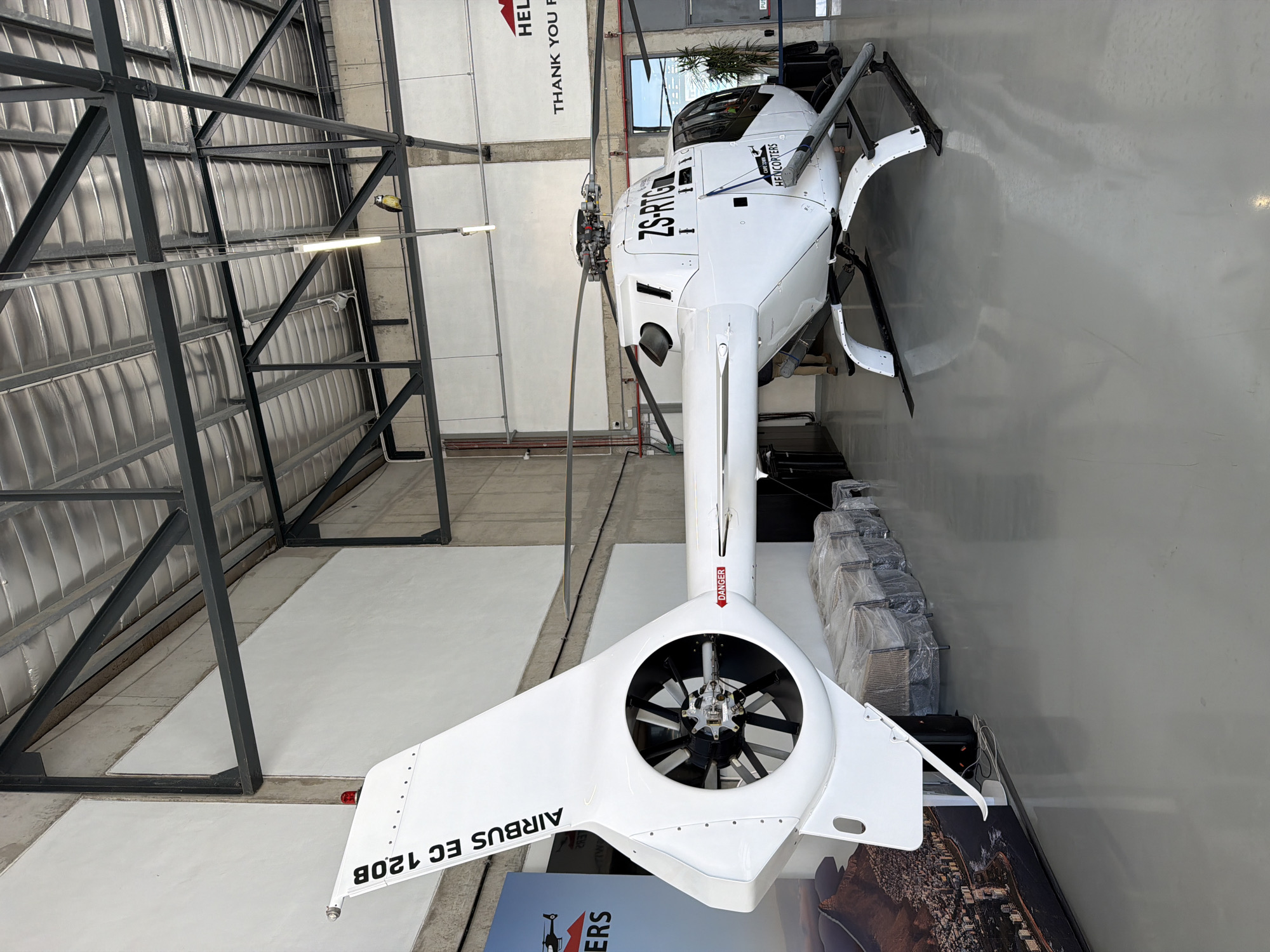 The image displays a white Airbus EC 120B helicopter parked indoors, likely within a hangar or exhibition space in Cape Town, South Africa. The helicopter, viewed from its rear-right, is the dominant subject, with its main rotor blades visible towards the front of the aircraft (top-left of the rotated image).
Notable text on the helicopter includes "ZS-RTG" on its fuselage, indicating a South African registration. The model "AIRBUS EC 120B" is printed on the tail fin, and a "DANGER" sign with an arrow is visible on the tail boom, pointing forward. The Fenestron (shrouded tail rotor) is clearly depicted at the very rear.
The setting features a high ceiling with exposed metal structural beams and corrugated panels, characteristic of an industrial building or hangar. Fluorescent light fixtures are visible. The floor is a polished, light-colored concrete. In the background, stacks of items, possibly equipment or seats, are covered in clear plastic sheeting. A large sign on the wall behind the helicopter's front section shows partial text such as "HELICOPTERS," "CAPE TOWN," "HEL," and "THANK YOU F," corroborating the provided location context of Cape Town. The helicopter appears to be stationary, possibly on display, in storage, or undergoing maintenance. There are no people visible in the scene.