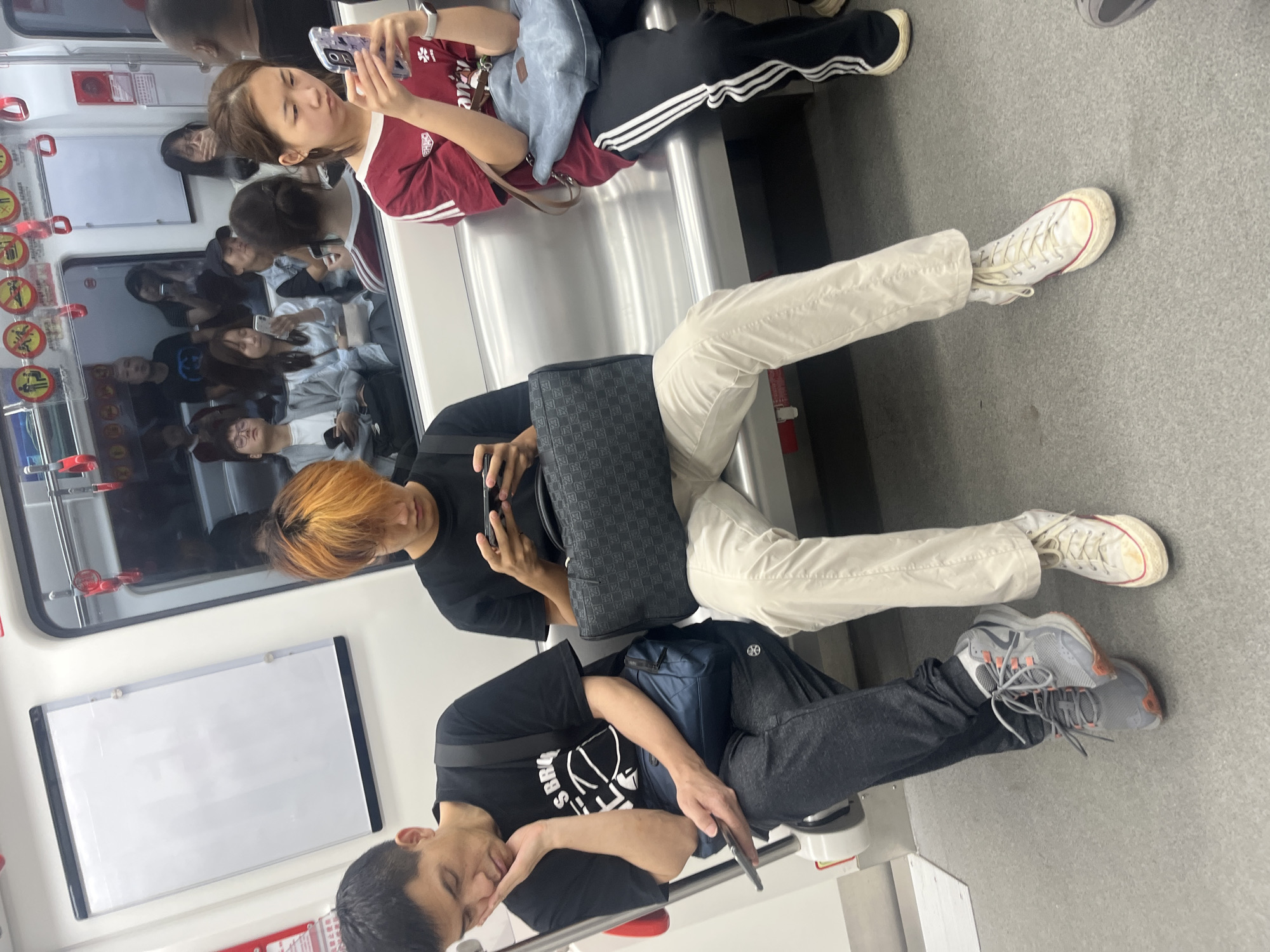 In an indoor subway train in Shapingba District, China, three individuals are seated on a metallic bench. On the left, a man with short black hair wears a black t-shirt with a graphic print and dark gray sweatpants. He rests his head on his hand, his eyes closed, holding a mobile phone in his right hand. Next to him, a person with dyed orange hair and a black t-shirt is seated, looking down at a mobile phone held in their hands. This person is wearing light-colored pants. To the right, a woman with dark hair pulled back wears a red t-shirt with white stripes on the sleeves and black athletic pants with white stripes. She is looking at her mobile phone, which she holds in her right hand. All three are positioned side-by-side on the bench, with the reflective train window behind them showing blurred images of other passengers. The environment is clean, with the floor appearing to be gray.