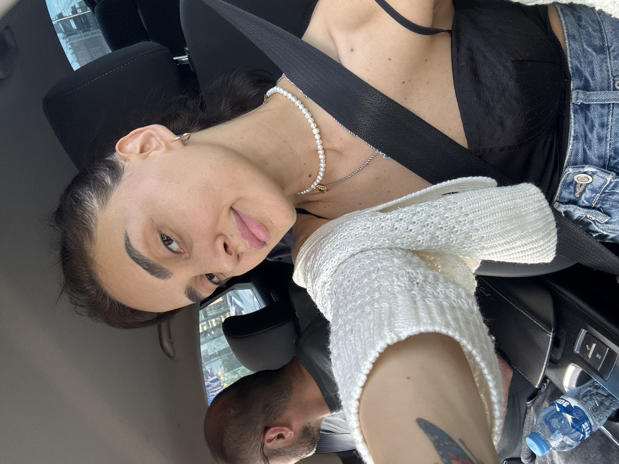 The image shows a woman in the passenger seat of a car, taking a selfie. She has dark hair pulled back and is wearing a white knitted cardigan over a black top and jeans. She is also wearing a pearl necklace and a silver necklace. A man with glasses is visible in the driver's seat, looking away from the camera. A bottle of water is in the center console of the car. The background outside the car windows appears to be a city scene with buildings, suggesting the car is likely in an urban environment, possibly Tbilisi, Georgia.
