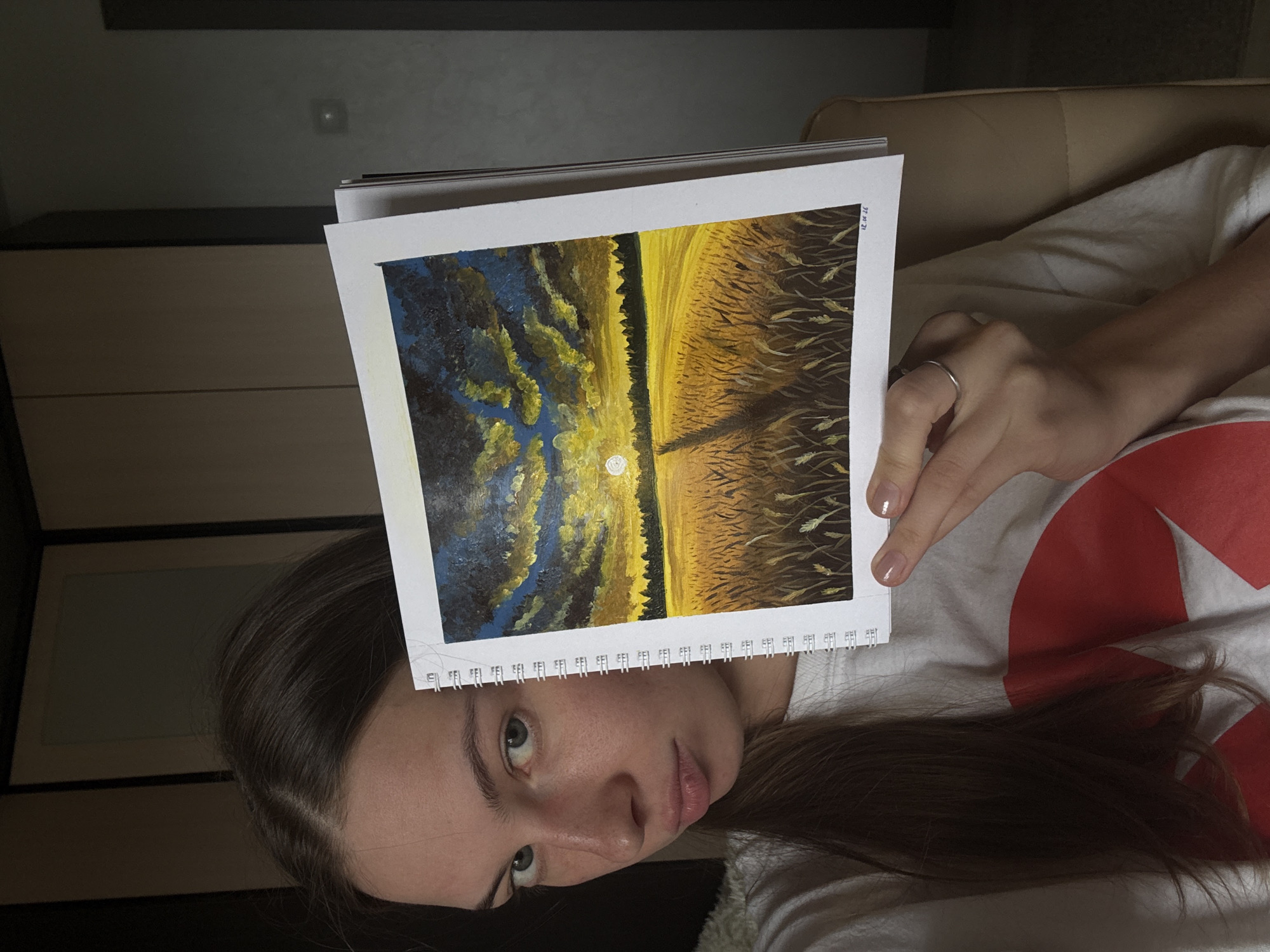 The image shows a young woman lying down, holding a painting above her face. The painting depicts a landscape with a field of wheat under a dramatic sky with clouds and the moon. The woman has long brown hair and is wearing a white t-shirt with a large red design. She is wearing a silver ring on her left ring finger. The painting appears to be done with oil paints, with visible brushstrokes and a textured surface. The lighting suggests it is daytime, possibly late afternoon or early evening, given the warm tones in the painting and the shadows in the room. The setting appears to be indoors, possibly a living room or bedroom, with furniture visible in the background. The woman's expression is neutral, and the overall mood of the image is relaxed and informal. There is some faint text in the upper right corner of the painting, but it is illegible. The location context is Perm, Russia.