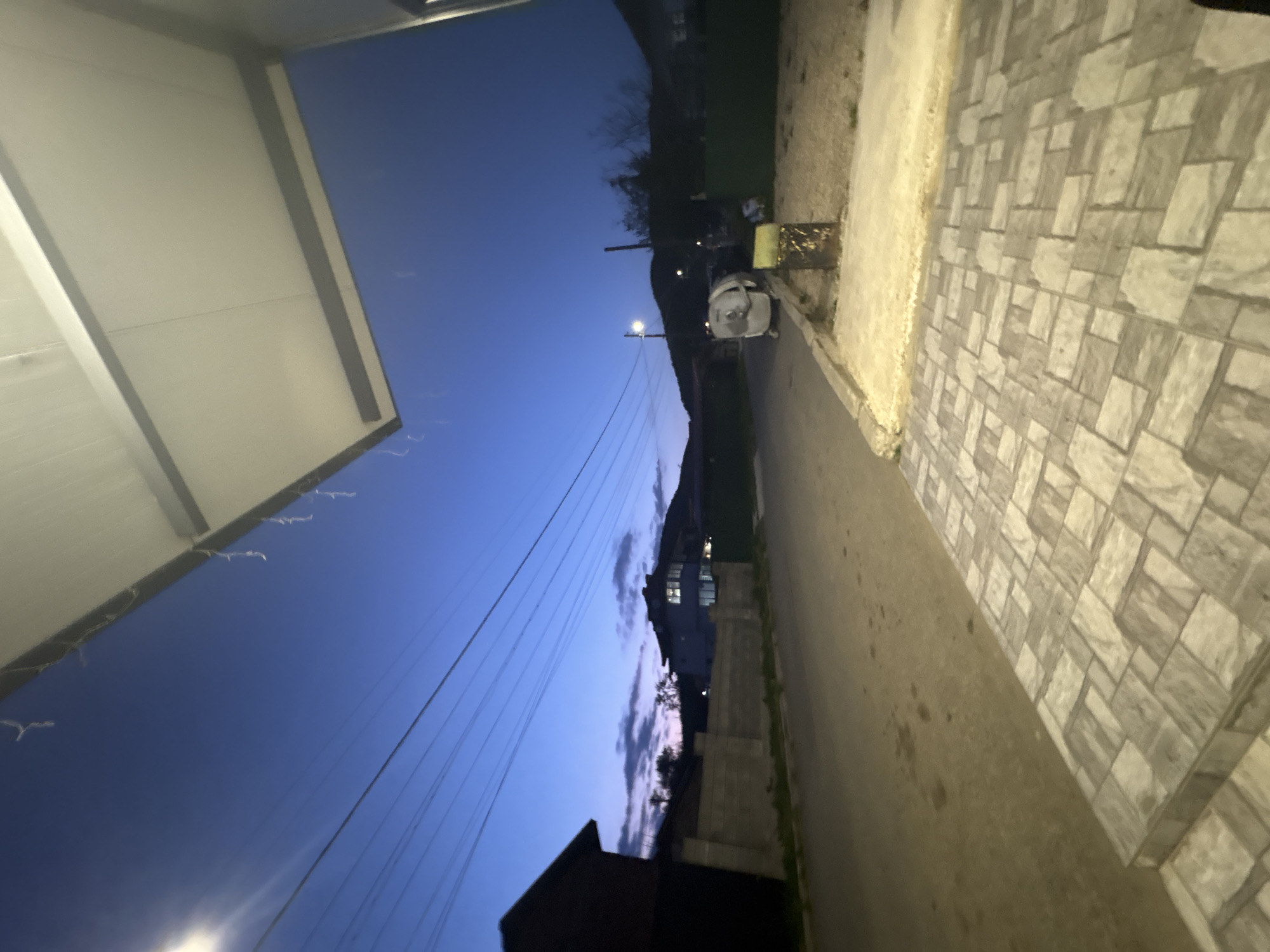 The image depicts a residential street in Goritsa, Bulgaria at dusk. The sky is a deep blue, fading to lighter hues near the horizon. Overhead power lines are visible. On the left, a structure with a bright, flat roof is present. A tiled sidewalk runs along the right side of the frame. Across the street, houses with illuminated windows can be seen, as well as trees and greenery. A street light illuminates a trash can on the right. A vehicle is parked near the trash can. The street appears to be empty of people.