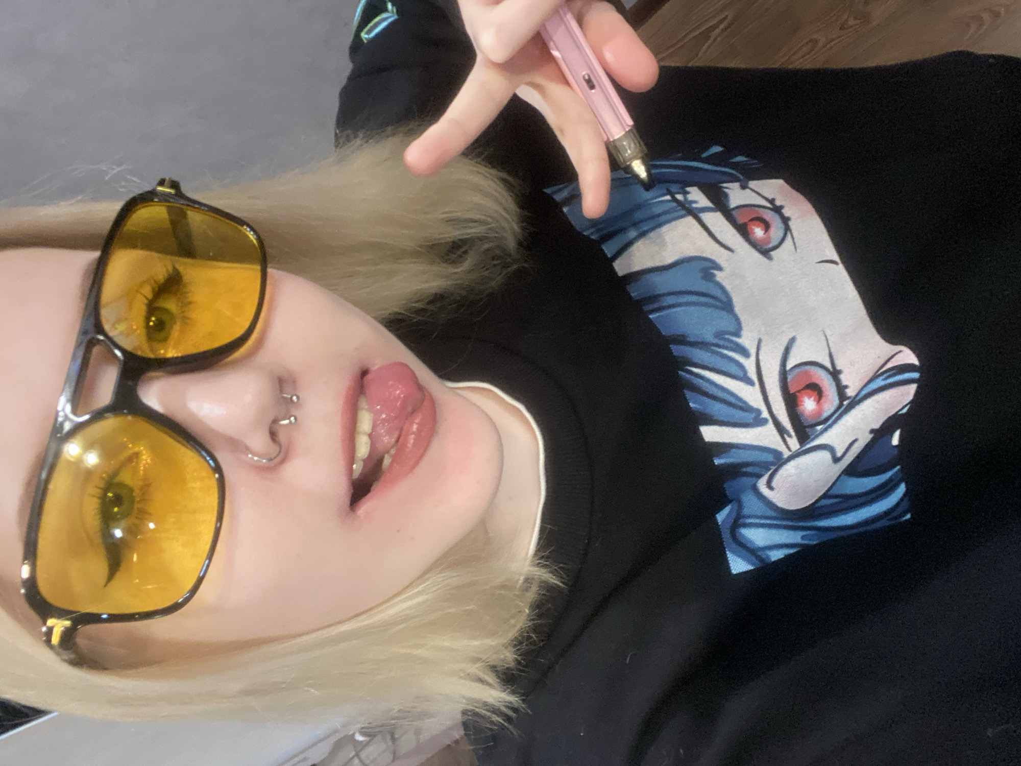 The image displays a close-up, frontal portrait of an individual, likely female, with blonde hair, captured indoors. The individual is wearing black-framed sunglasses with yellow-tinted lenses, and dark eyeliner is visible beneath them. The nose features two piercings: a silver septum horseshoe barbell and a silver nostril ring on the viewer's left. The mouth is open, with the tongue extended. The individual wears a black long-sleeved garment with a thin white collar trim, featuring a large graphic print of an anime-style character's blue hair and red eyes on the front. The individual's left hand, positioned on the viewer's right, forms a "peace" sign with the index and middle fingers extended. A pink electronic vaping device is held between these fingers and the thumb. The background consists of a plain, light-colored surface on the left and a darker, possibly gray, wall on the right, which transitions to a wooden surface at the bottom right. No visual elements within the image confirm the specified location of Izhevsk, Russia.