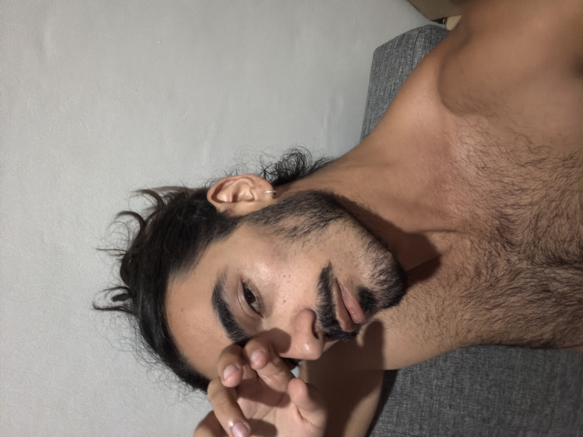 The image, rotated counter-clockwise, captures a close-up of a man lying on his left side, looking directly at the viewer. He has dark, curly hair, a full beard, and a mustache. His expression is calm and somewhat pensive, with his lips slightly parted. His left hand is brought up towards his face, with his fingers resting near his mouth and nose area. A small silver hoop earring is visible in his left ear.
The man is shirtless, revealing his chest and shoulder, which are covered in dark body hair. He is resting against a dark grey, textured fabric surface, likely a pillow or a part of a couch or bed. The background is a light grey, textured wall, creating a simple indoor setting. The lighting is soft, casting subtle shadows that highlight his facial features and the contours of his body. There are no discernible location cues specific to North Kuta, Indonesia, visible in the image itself, nor is any text present.