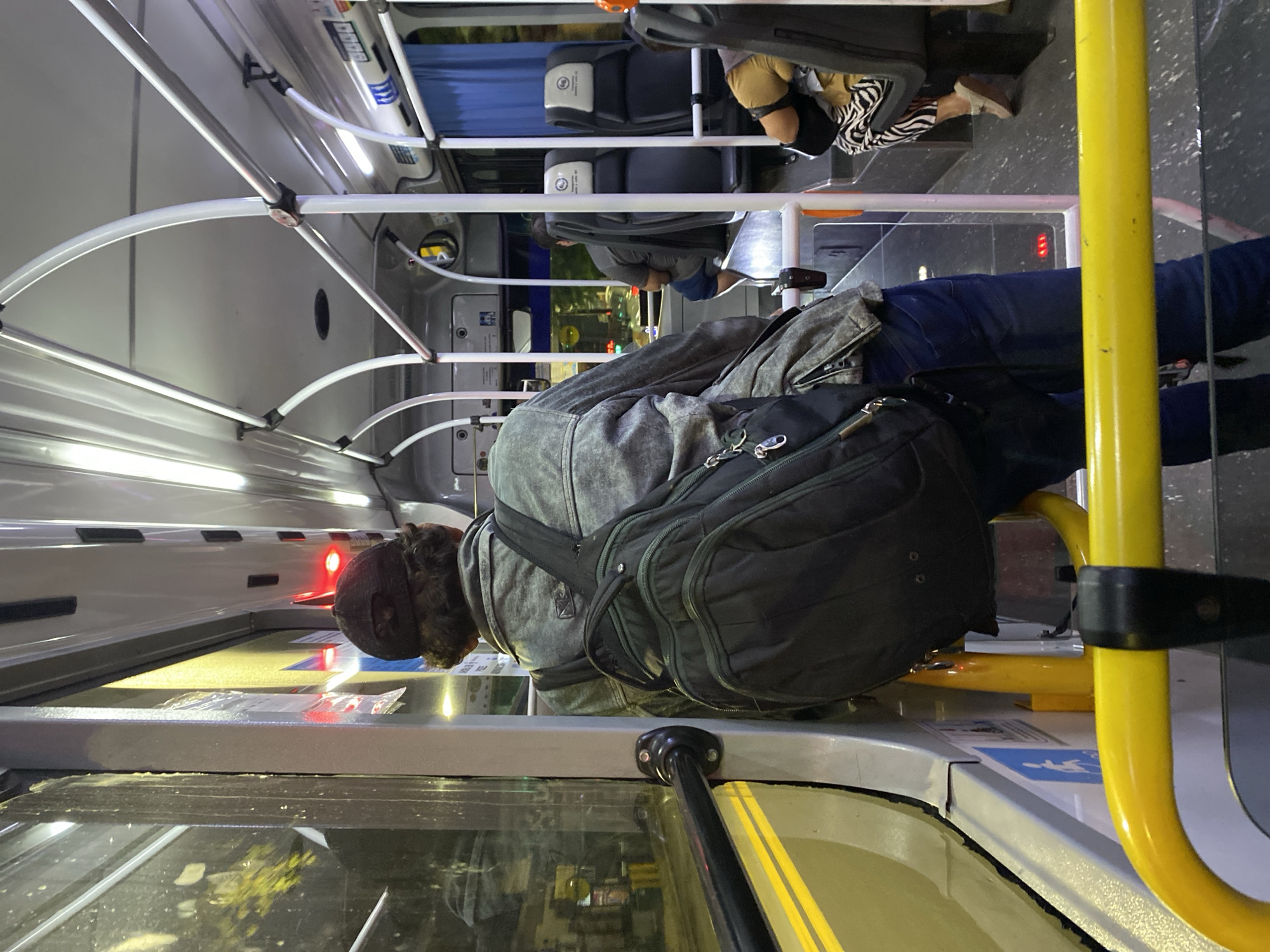 The image captures a scene inside a bus. A person wearing a grey jacket and a dark backpack stands, holding onto a yellow pole. Other passengers are seated in the background. Through the bus window, a blurred cityscape is visible, suggesting an urban setting. The interior of the bus is brightly lit. The location is likely Buenos Aires, Argentina.