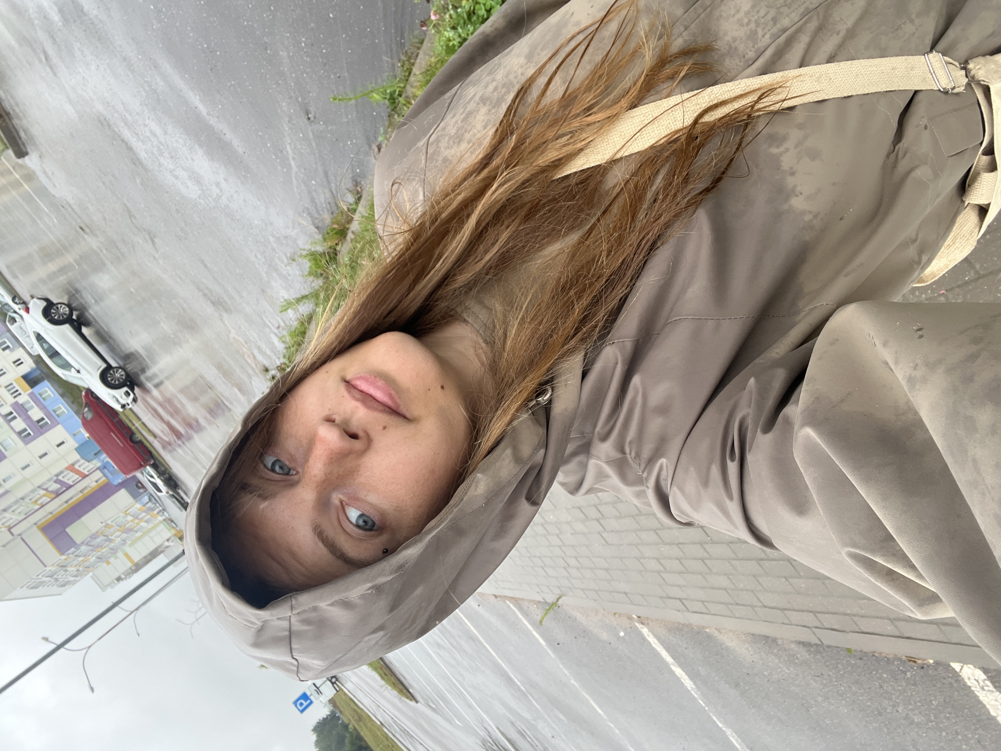 A woman with long, brown hair, wearing a tan hooded jacket and a light-colored strap across her chest, takes a selfie outdoors on a cloudy day in a wet, paved area. The background shows a parking lot with several cars, a red van, and a multi-story building with colorful panels. The ground is wet, indicating recent rain. The woman has light blue eyes and a few freckles on her face. The scene is located on the territory of the Sennitsy rural council in Belarus.