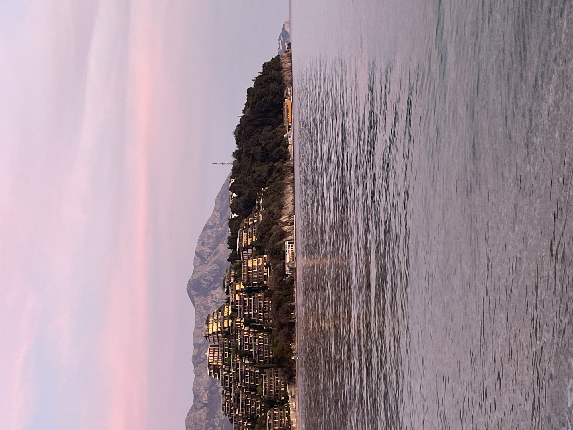 The image displays a coastal landscape featuring Sveti Stefan, Budva, Montenegro, during twilight. The sky, occupying the upper third, presents a gradient of light pink and lavender hues. In the background, rugged grey mountains are visible. The midground is dominated by a densely forested peninsula extending into calm water, which shows subtle ripples and reflections. A prominent complex of multi-tiered, terraced residential or hotel structures with balconies is built into the hillside of this peninsula, illuminated by a warm, ambient light. To the right, atop the forested section of the peninsula, a red and white lattice tower is visible. Along the water's edge on the right side of the peninsula, a long, low-profile yellow and white vessel or structure is docked. The foreground consists entirely of the calm, reflective water. No human figures or discernible actions are present in the scene.
