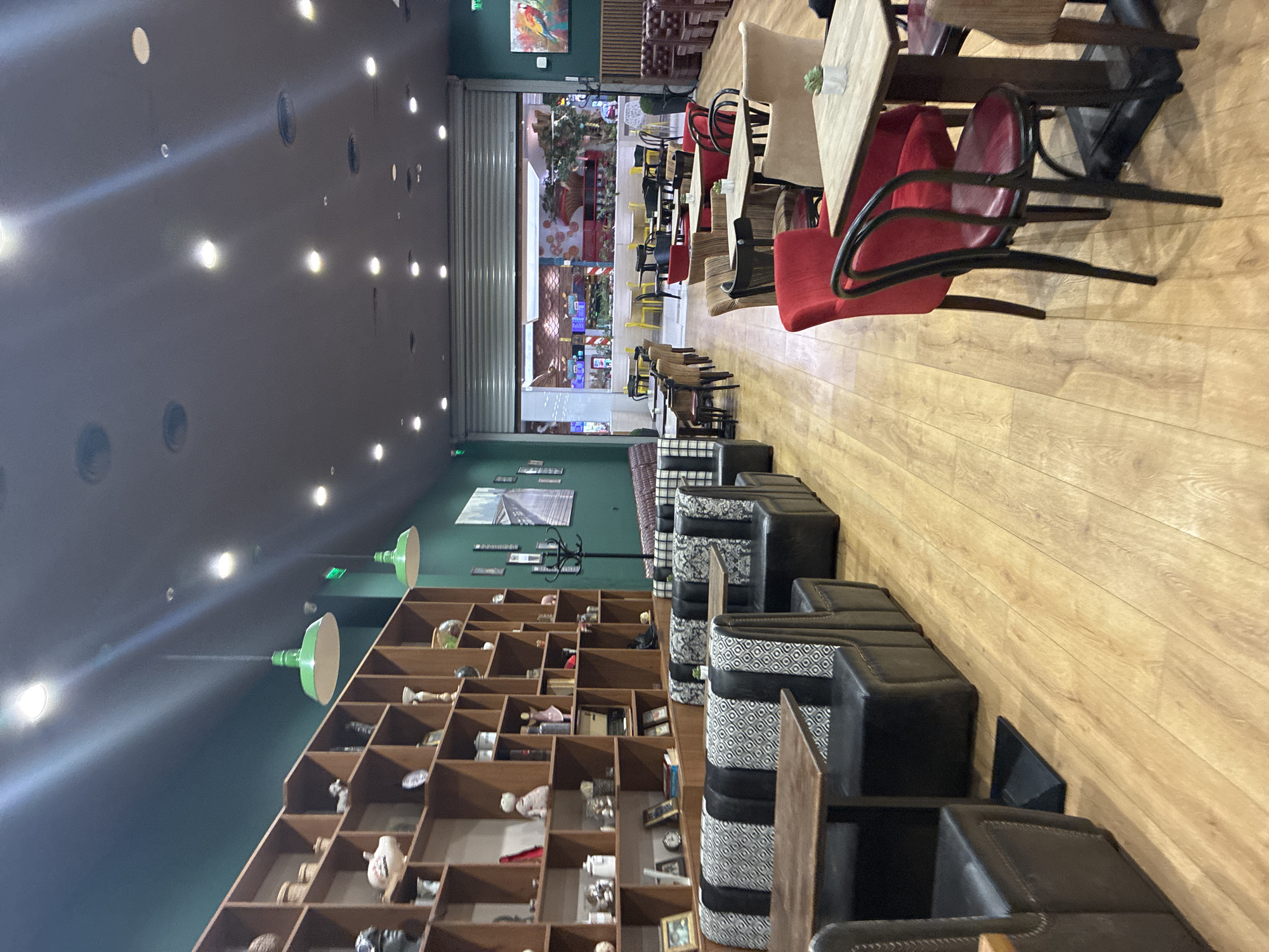 The image shows the interior of a restaurant in Plovdiv, Bulgaria. The scene is captured from a low vantage point. A large shelving unit filled with various objects lines the left wall. The wall is painted in a dark green color. The ceiling is dark and features recessed lighting. Tables and chairs are visible on the right side of the image. The floors are wooden. At the back of the restaurant, there's a view to what appears to be an external shopping area with decorations. A row of booths with patterned fabric and dark leather seats runs alongside the shelves. There are no people visible in the immediate foreground.