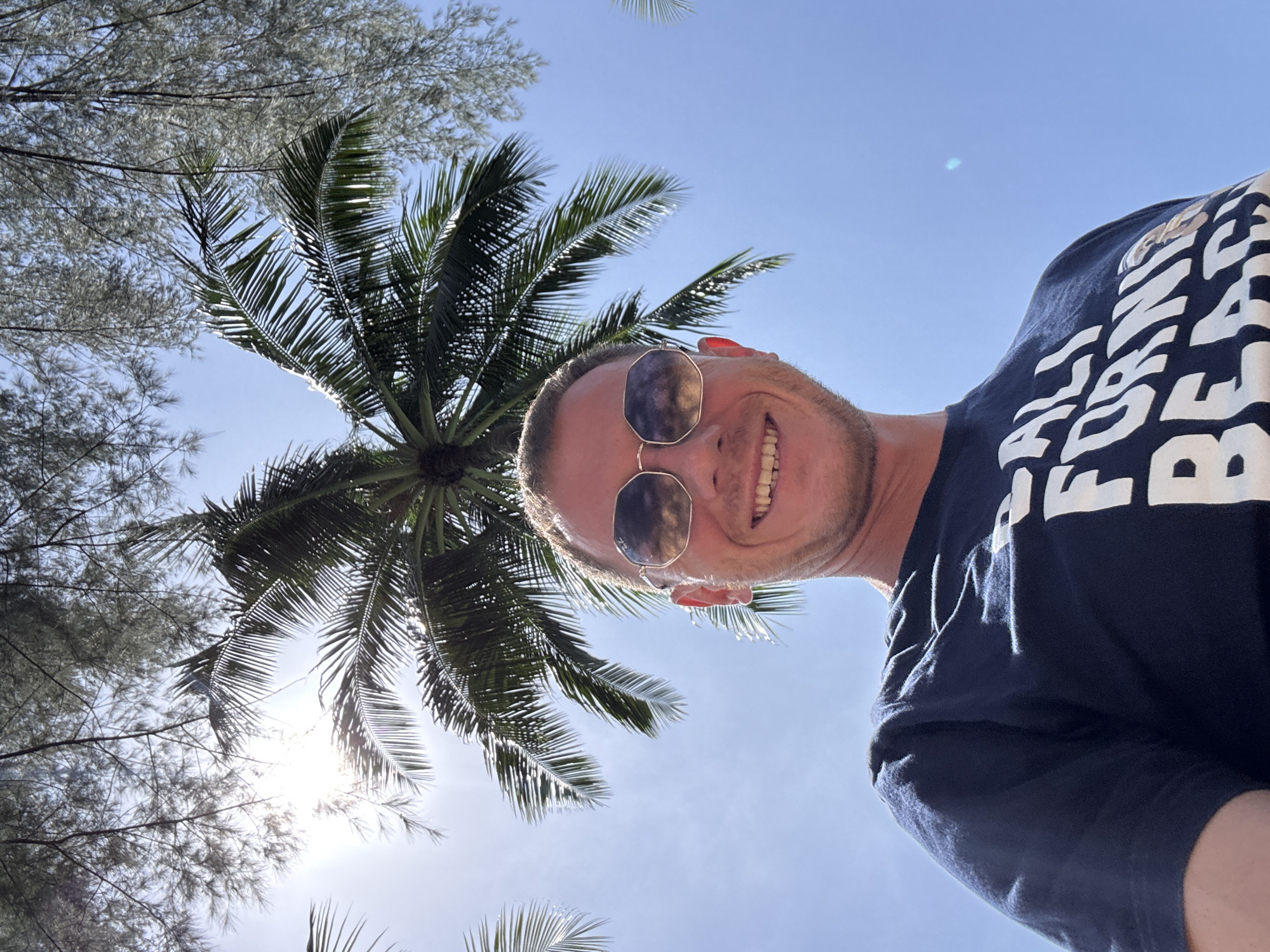 A man with sunglasses and a navy blue t-shirt is captured in a selfie outdoors. He is smiling and looking at the camera. The t-shirt has white lettering that says "CALI FORNIA BEACH" along with an image of a sloth. The man is positioned in the foreground, with palm trees and the sky visible in the background. The sun is bright, casting a glare and some shadows. The scene appears to be a sunny day, possibly at a beach location. The location is identified as Ban Khuk Khak, Thailand.