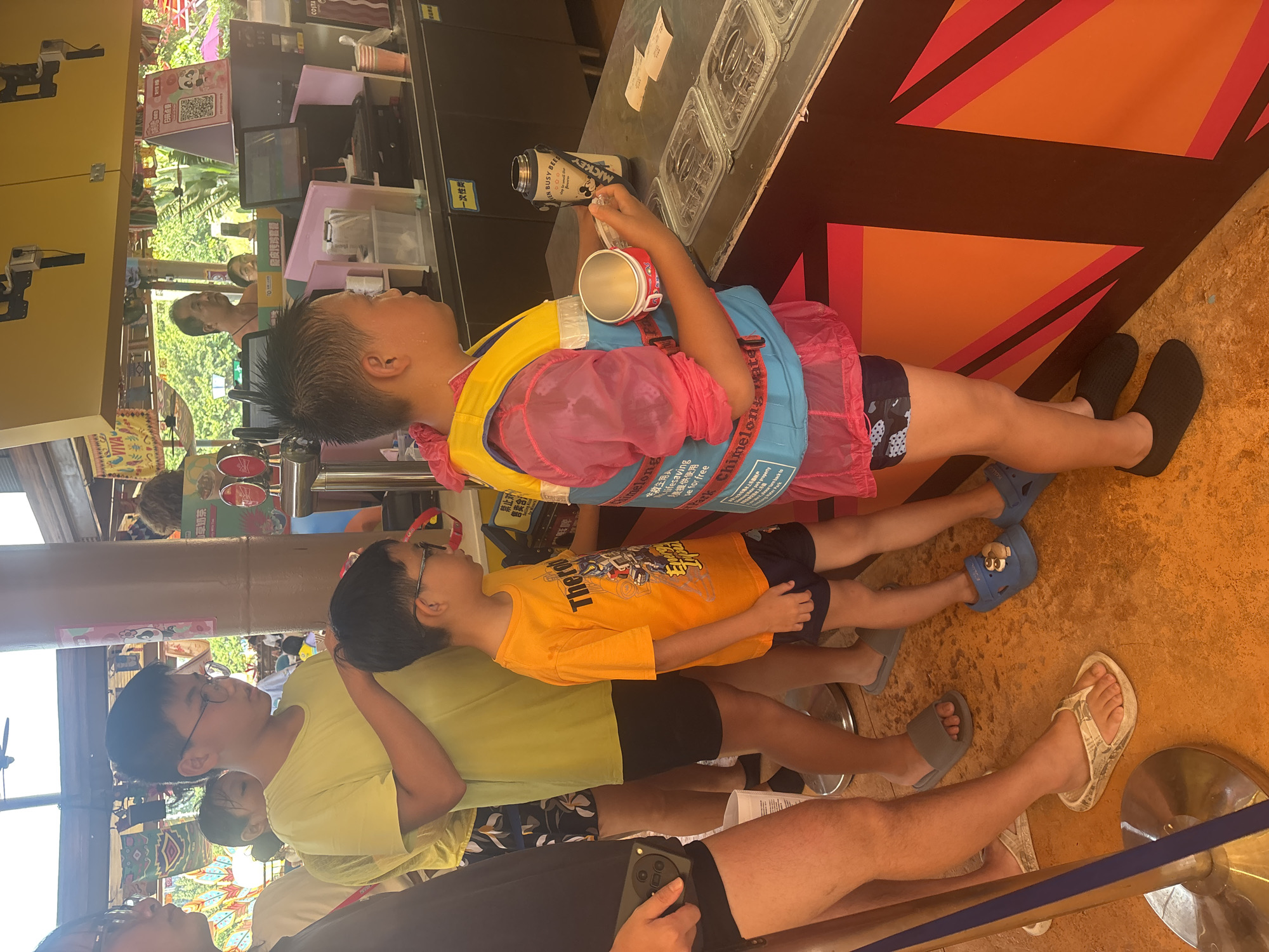 In Panyu District, China, a group of children and adults are queuing at a service counter within what appears to be an amusement park or water park. The counter is part of a building with a yellow awning overhead, and a tropical-themed backdrop with palm trees is visible in the distance. The flooring is a textured orange surface, and a blue stanchion is positioned in the foreground.
The individuals are primarily children, dressed in casual summer attire. One boy in the center wears a yellow t-shirt and dark shorts, with glasses and dark hair. To his right, another child is dressed in a vibrant pink and blue life vest over a blue and black patterned swimsuit. This child holds a white metal cup and appears to be interacting with the counter. Another child, wearing an orange t-shirt with a graphic design, stands next to the boy in the yellow t-shirt. Adults are also present, with a woman in a black t-shirt and patterned shorts visible on the left, and another adult further in the background. The counter itself has a dark countertop with several clear plastic containers arranged on it. Behind the counter, a beverage dispenser and a cash register system are visible. Promotional posters and signage are also present in the background, with one poster featuring a QR code and a cartoon panda character.
