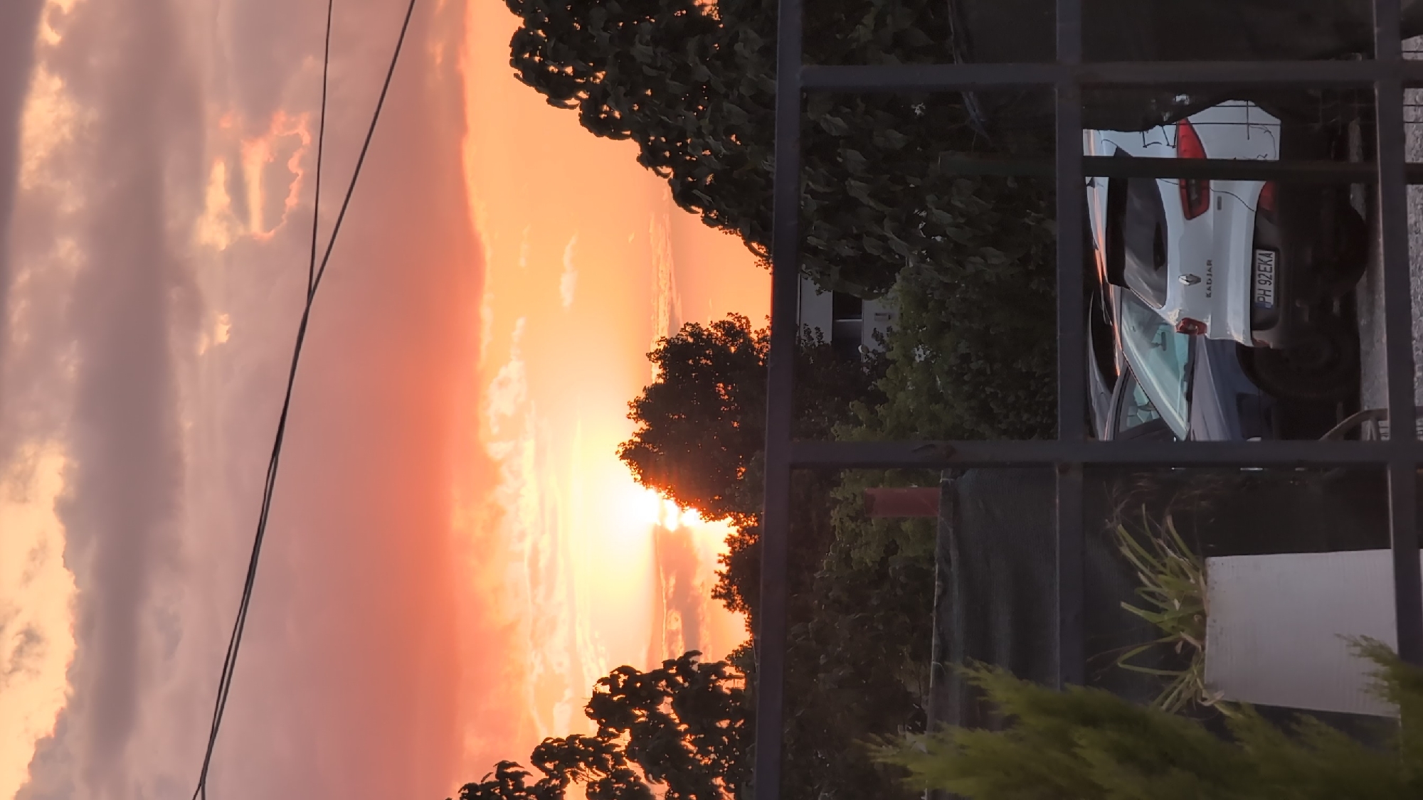 The image captures a dramatic sunset over what appears to be a residential or semi-urban area in Constanța, Romania. The sky is ablaze with vibrant hues of orange, pink, and purple, with clouds catching the last rays of the sun. The sun itself is low on the horizon, casting a warm glow and creating a silhouette effect for the trees in the foreground.
The scene is framed by a dark, metal fence or railing, which partially obscures the view of parked cars and some foliage on the right side of the image. One of the cars, a white vehicle, has "KADJAR" written on its rear. The license plate is visible and reads "PH 92EKA".
The lighting suggests that it is either early morning or late evening, with the sunset being the dominant feature. The weather appears to be clear, allowing for the spectacular display of colors in the sky. There are no people visible in the image. The overall mood conveyed is one of peacefulness and natural beauty, amplified by the striking sunset. Power lines are also visible, stretching across the sky.