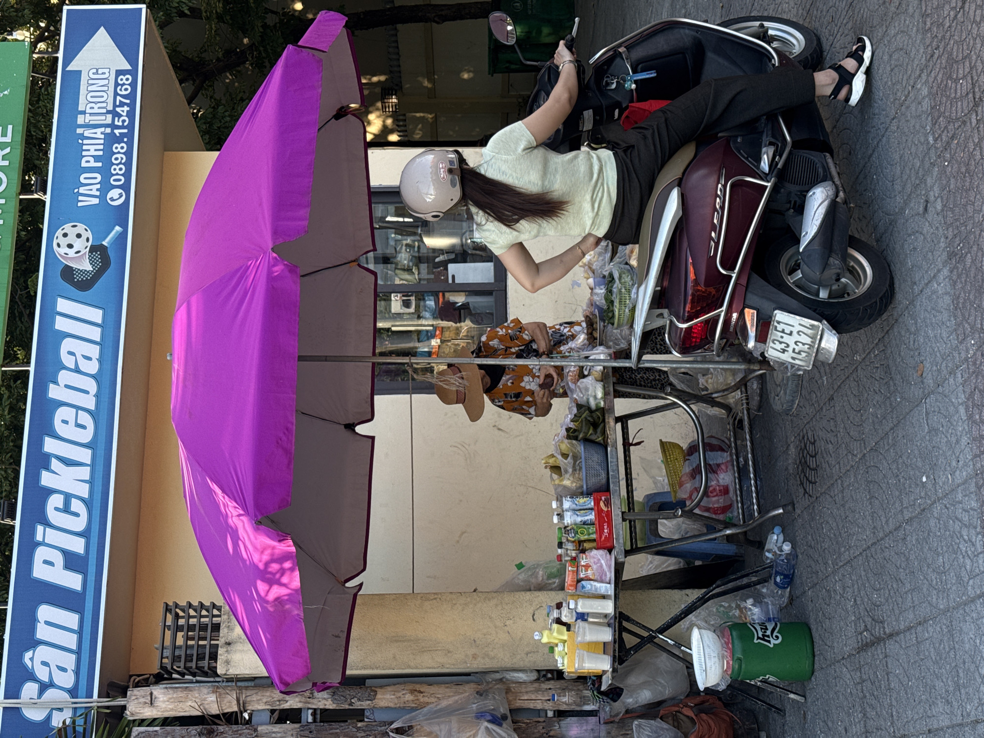 The image depicts a street scene in Da Nang, Vietnam. A woman wearing a light green short-sleeved shirt, dark pants, and sandals is seated on a dark red scooter, facing away from the camera. She has long dark hair and is wearing a light-colored helmet. Her left hand is on her hip, and her right hand is near the scooter's handlebars. In front of the scooter, a table is laden with various items, including bottled drinks, plastic cups, and bags of produce. A woman wearing a hat and an orange patterned shirt is standing behind the table, appearing to be tending to the merchandise. A large purple umbrella is providing shade over the table and the area around it. The ground is paved with textured concrete tiles. A sign above the scene reads "San Pickleball" and "Vào Phía Trong" with a phone number. The general environment suggests a bustling street market or roadside stall.