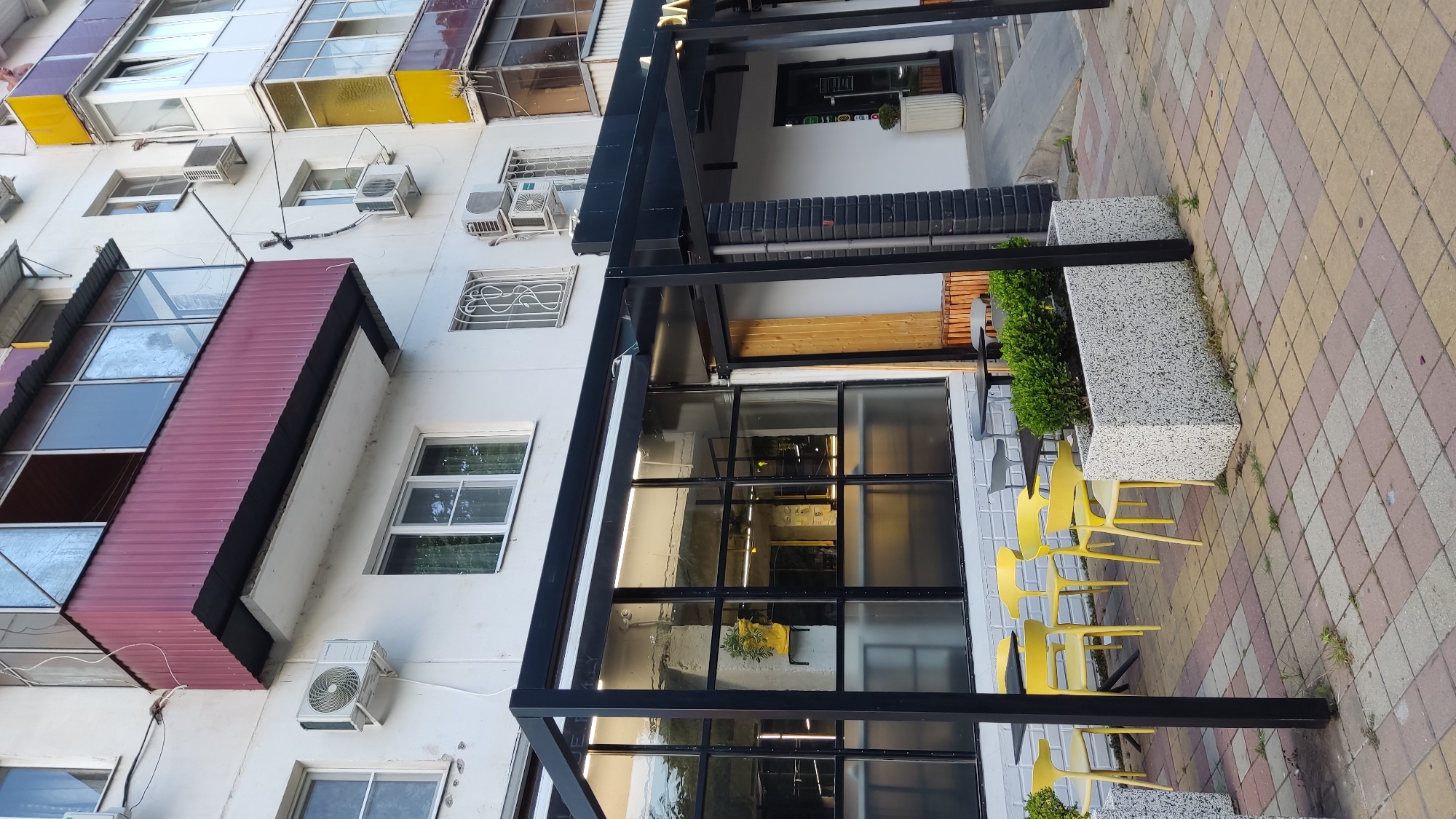 The image depicts a street-level view of a modern building with an outdoor seating area for a cafe or restaurant. The building has a light-colored facade with multiple windows and balconies. The ground floor features a large glass-fronted area, likely the interior of the establishment, with visible lighting and decor including a yellow vase on a pedestal. An awning extends over this section, supported by black metal poles that also frame the entrance.
Outside the glass enclosure, there are several tables with yellow chairs arranged on a patterned tiled sidewalk. Adjacent to the seating area are concrete planters filled with greenery. The sidewalk itself is paved with square tiles in a pattern of yellow, pink, and beige. The building appears to be located in a city, with elements like air conditioning units mounted on the facade and windows suggesting residential or commercial units above. The specific location, as provided, is Krasnodar, Russia. No people are clearly visible in the image, and there are no apparent actions or interactions taking place. The scene is static and presents an exterior view of a commercial establishment on a street.