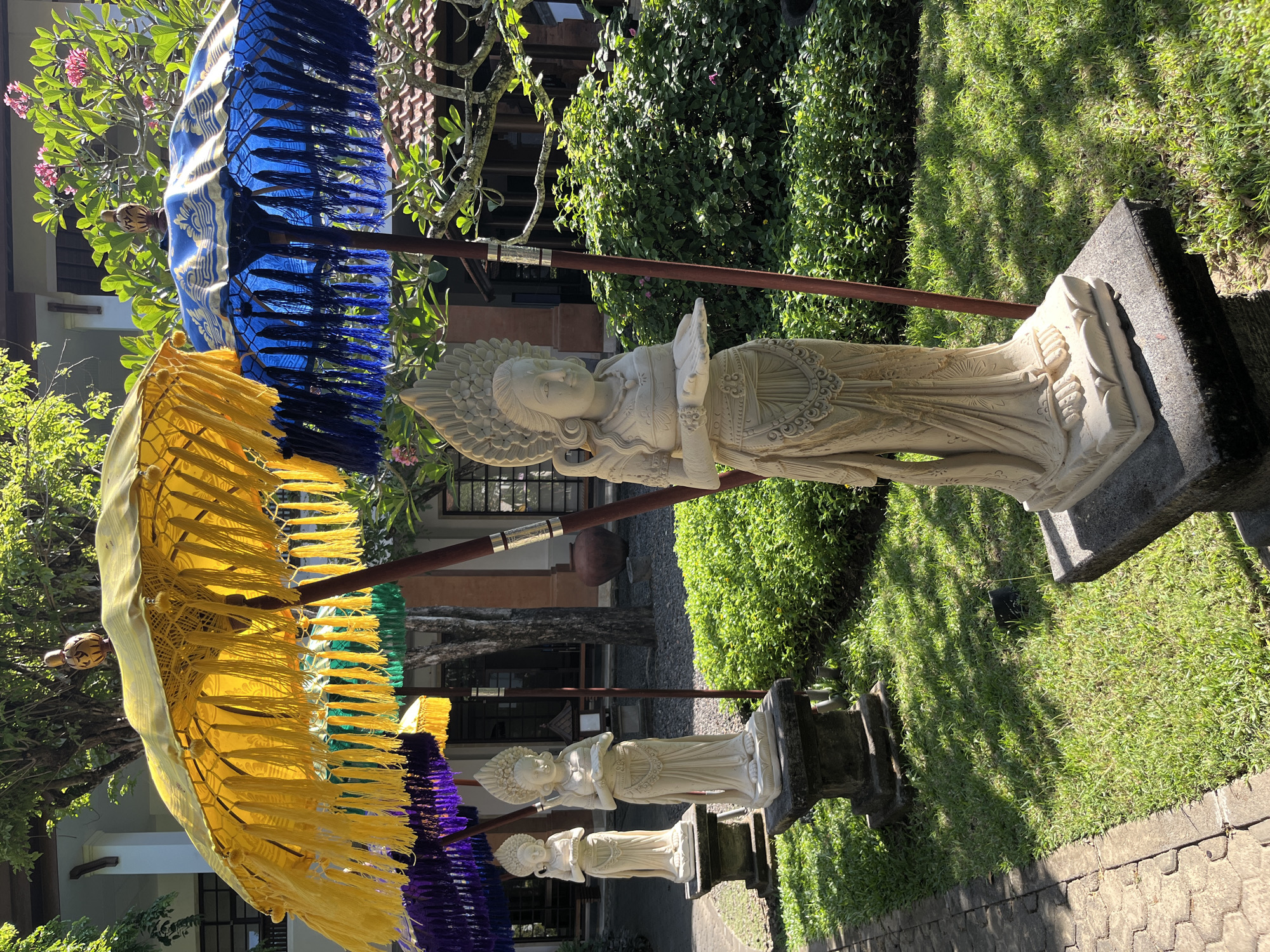 The image depicts a sunny courtyard in Kuta Selatan, Indonesia. In the foreground, a light-colored stone statue of a woman with intricate headdress and flowing garments stands on a pedestal. To the left and behind her, two other similar statues are visible. They are adorned with decorative umbrellas in vibrant colors, including yellow, blue, purple, and green, each with hanging tassels. The statues are placed on stone bases set against a backdrop of lush green grass and dense shrubbery. In the background, part of a building with tiled roofs and windows is visible, with trees and foliage casting shadows on the ground. The scene suggests an outdoor area, possibly a garden or a temple complex.
