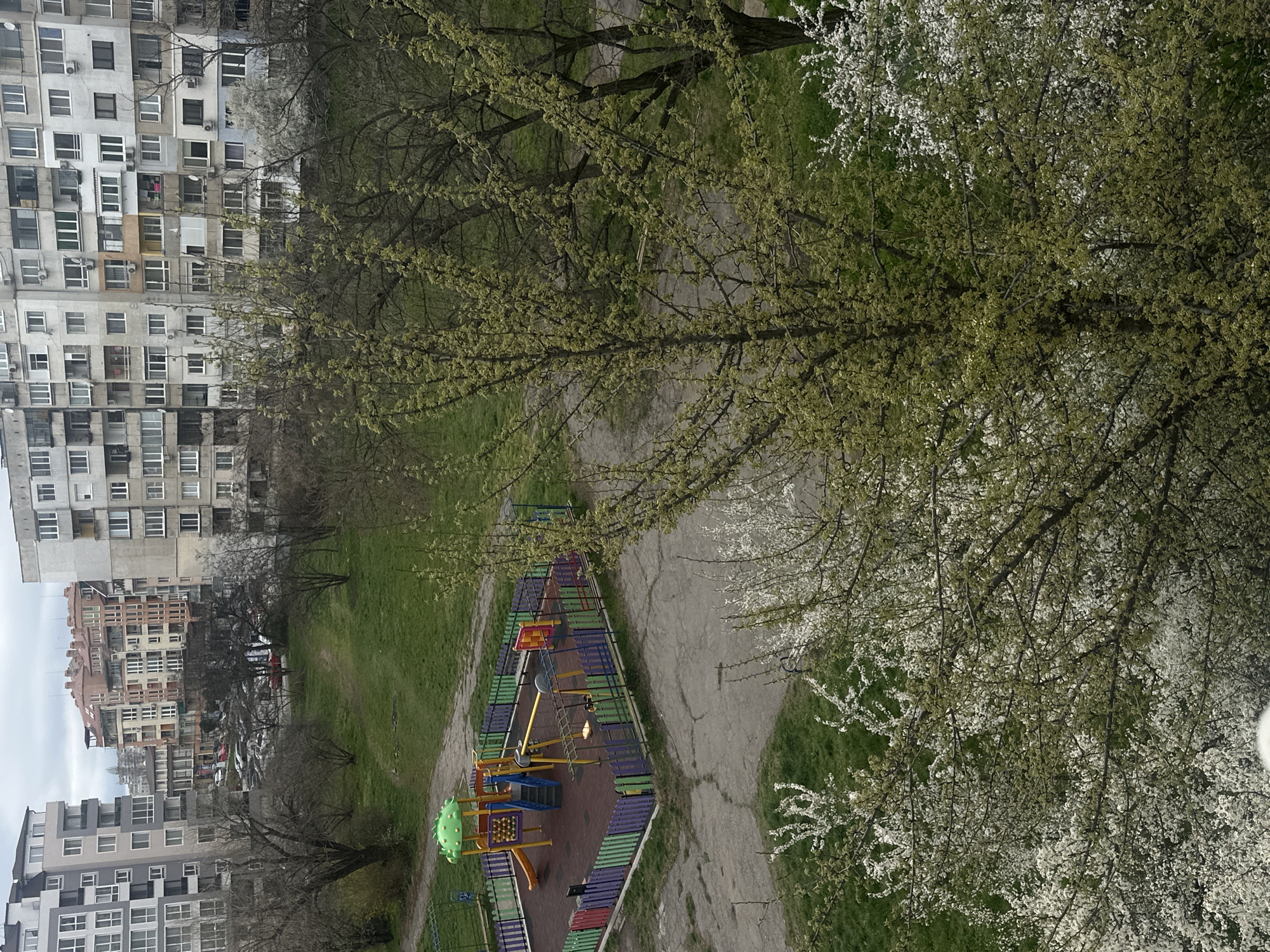 The image shows a playground in Sofia, Bulgaria, viewed from above. The playground has a colorful perimeter fence and includes equipment such as a slide, climbing structures, and a seesaw. A cracked concrete path leads to and from the playground. The area around the playground is grassy with several trees, some of which are in bloom with white flowers. Apartment buildings are visible in the background, indicating an urban residential area. The sky is overcast.