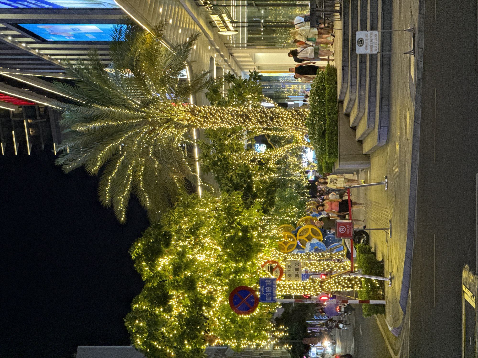 The image depicts a nighttime scene on a city street, likely in Nha Trang, Vietnam, adorned with festive yellow lights. Trees lining the sidewalk are densely wrapped in strings of warm white lights, creating a luminous canopy. A tall palm tree, also illuminated, stands prominently in the foreground, its fronds outlined with lights.
On the sidewalk, several individuals are visible. A group of people, including men and women dressed in casual attire, are walking or standing. Some are looking towards the entrance of a modern building that appears to be a commercial establishment or hotel, characterized by its sleek facade and illuminated signage. Steps lead up to the building's entrance.
Several street signs are present. One is a circular blue and red sign with a horizontal line and a cross, indicating a restriction. Another sign, in Vietnamese, provides information. Decorative elements, including yellow and blue circular motifs, are also placed along the sidewalk.
A street runs alongside the decorated trees, with a few motorcycles visible in the background. The overall atmosphere is one of evening activity and festive illumination in an urban setting.