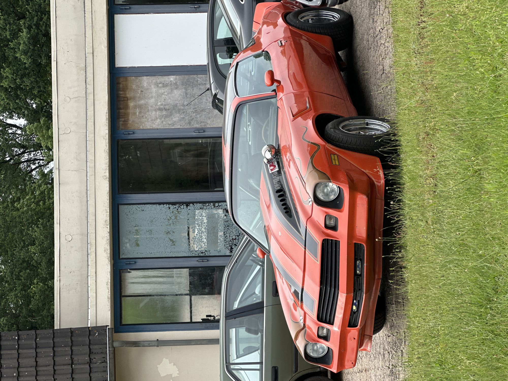 An outdoor image, captured in Kutenholz, Germany, presents a classic orange American muscle car, likely a second-generation Chevrolet Camaro, parked on an asphalt surface. The car features bold black and brown/tan racing stripes and graphics extending across its hood and sides. A prominent chrome hood scoop with visible engine components bears the word "OUTLAW" in black text. The vehicle is equipped with polished chrome wheels and features T-top roof panels, with the driver's side visible.
Partially visible behind the orange car is a darker, modern vehicle, possibly a gray or black sedan. The background consists of a multi-story building with large, rectangular windows, some of which appear discolored, aged, or dirty. Beyond the building, lush green trees are visible. The foreground and right side of the image show a strip of green grass, indicative of an unmaintained area beside the cracked asphalt parking surface. The overall lighting suggests a bright, possibly overcast, daytime setting.