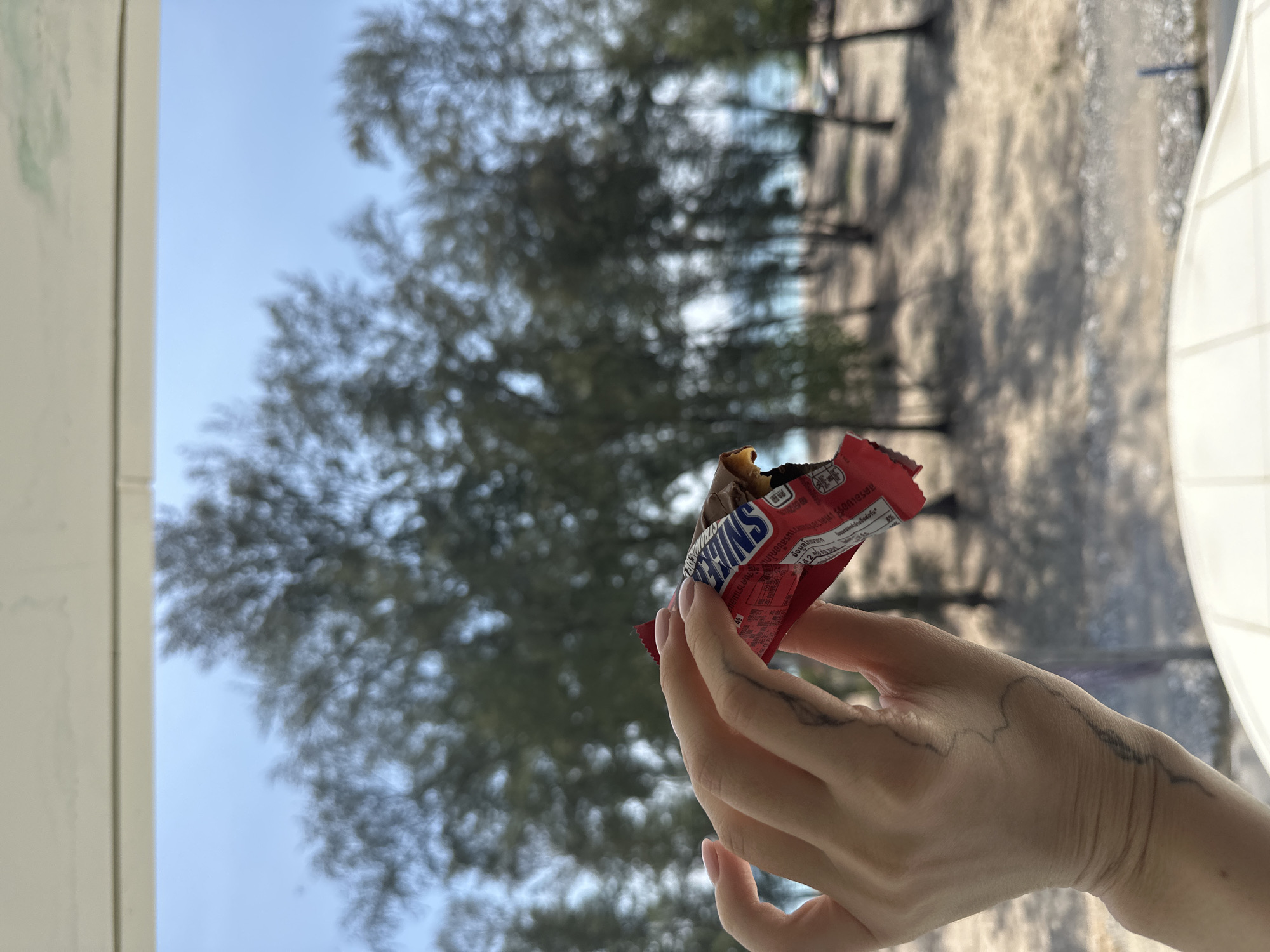 The image depicts a close-up view of a human left hand holding a partially opened Snickers candy bar, set against a blurred coastal landscape in Choeng Thale, Thailand. The hand has light-toned skin, with light pink nail polish visible on the fingernails. A dark, linear tattoo, resembling a geographical contour or map, is present on the back of the hand and extends onto the wrist. The candy bar wrapper is red and displays "Snickers" along with Thai script. The chocolate bar inside is partially consumed, revealing its nougat and caramel layers. The blurred background features a clear, light blue sky above a dense arrangement of tall, dark-foliaged trees, characteristic of coastal pine or similar vegetation. Beyond the trees, a sandy area transitions into a distinct light blue body of water, indicating the sea. A white architectural element is visible horizontally at the top of the frame, and a white tiled surface appears in the bottom right, suggesting the vantage point is from an elevated indoor or balcony setting.