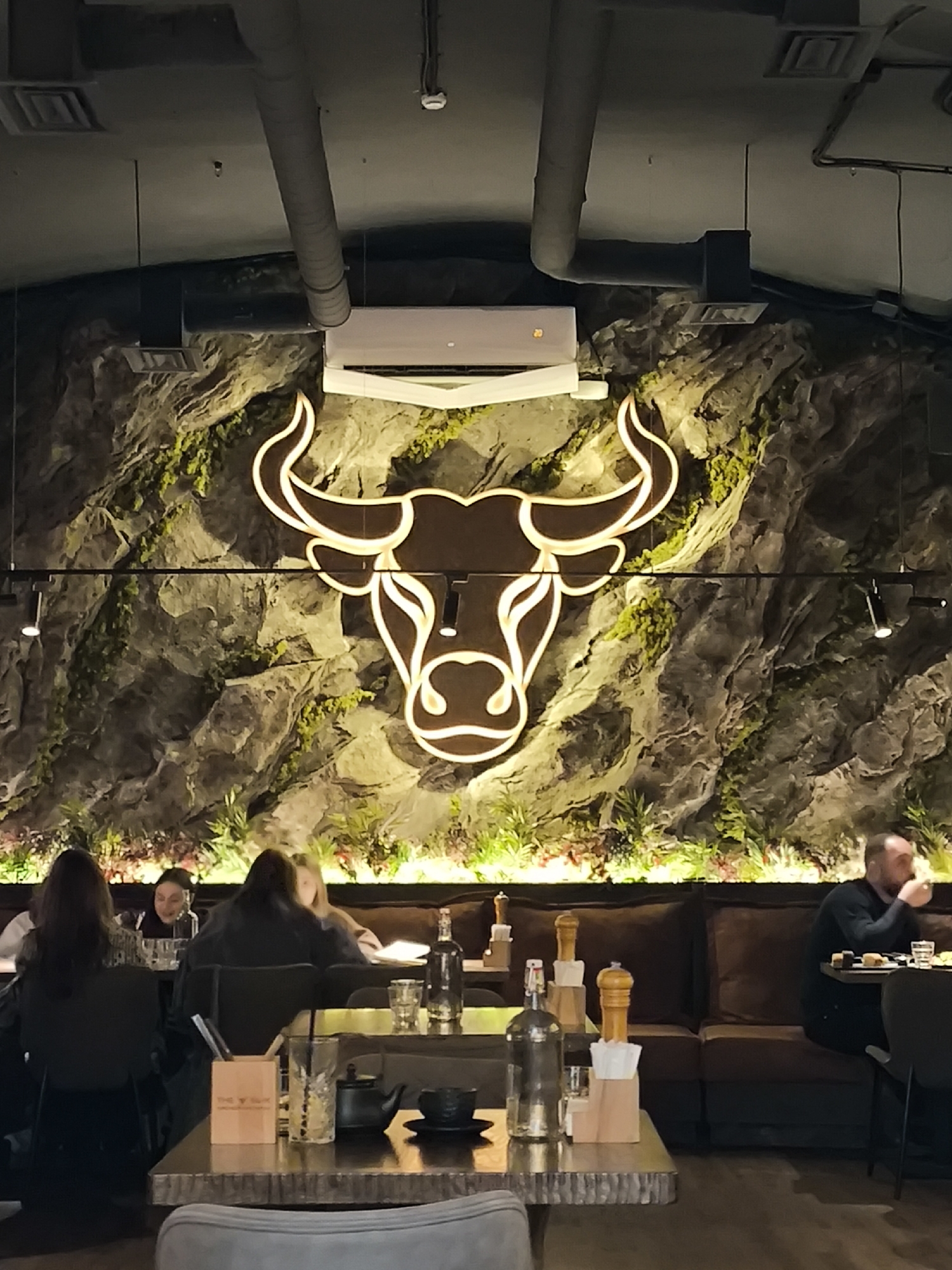 The image shows the interior of a restaurant in Unknown, Unknown. The focal point is a large, illuminated bull head logo mounted on a wall designed to resemble a rock face with moss and vegetation. The restaurant's ceiling is dark and features exposed air conditioning ducts and ventilation equipment. Patrons are seated at tables in the foreground, some occupied and some unoccupied. On one table, a tea set, drinking glasses, condiments, and a utensil holder are visible. In the background, a cushioned seating area runs along the wall. A man sits at one of the tables in that area, eating. Other diners are seated at tables in the left of the image. The lighting is subdued, creating a cozy atmosphere.