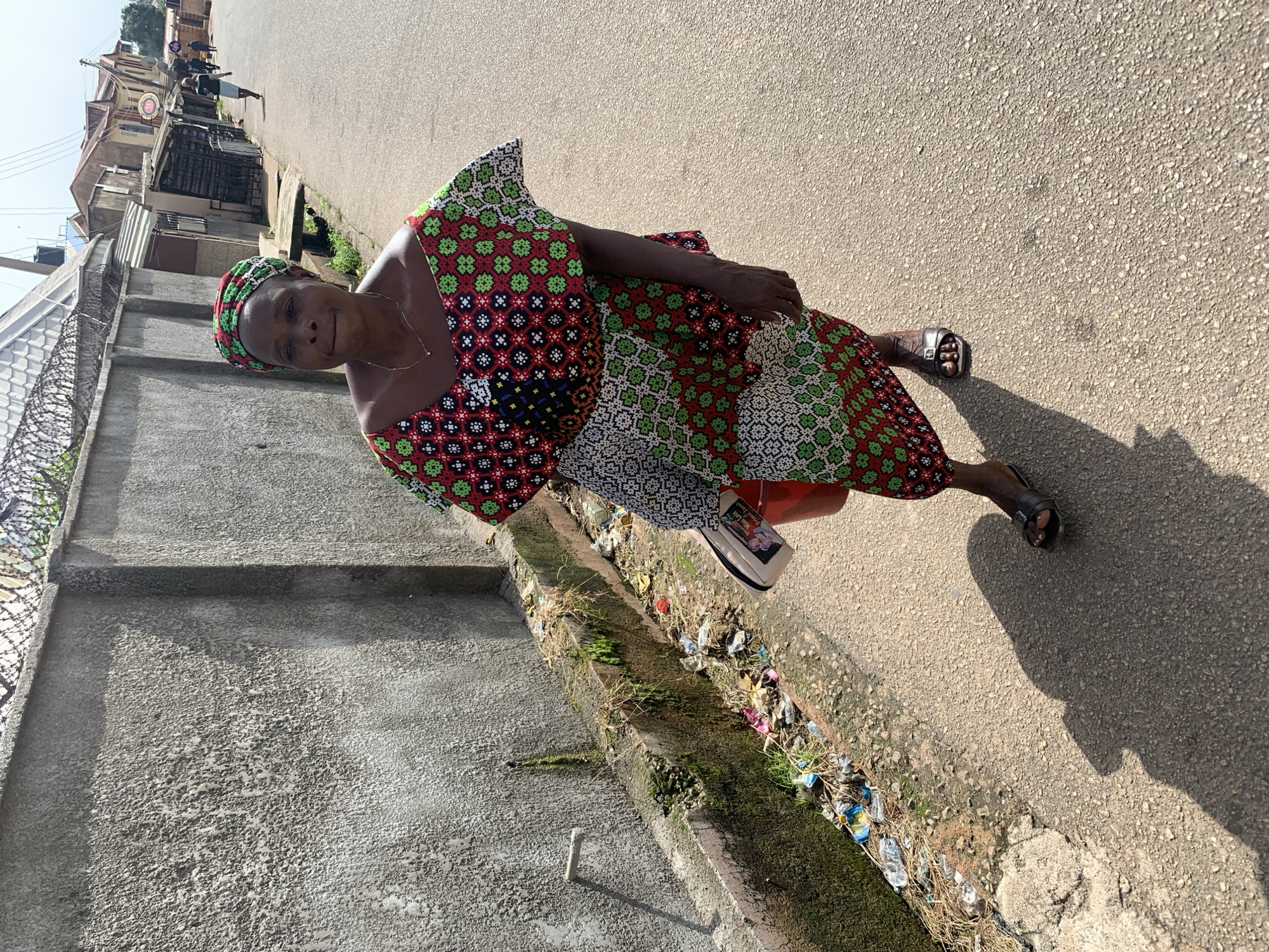 A Black woman is walking on a paved road in Jos, Nigeria, casting a shadow on the ground. She is wearing a colorful patterned headwrap and a matching dress with a V-neckline and short, flared sleeves. The dress is predominantly red and green with black and white geometric patterns. She is carrying a red bucket in her left hand and a light-colored purse with a picture on it in her right hand. She has dark skin and is wearing dark sandals.
The environment appears to be an urban street. To her left is a concrete wall with a rough texture, topped with barbed wire fencing. A narrow, overgrown drainage ditch runs along the base of the wall, filled with trash, debris, and some greenery. Beyond the wall, there are buildings with corrugated metal roofs and a few people walking in the distance. The street itself is paved and appears to be in decent condition, with some small pebbles and texture visible. The lighting suggests it is daytime, with a strong light source creating distinct shadows. The overall impression is of a busy, somewhat informal urban setting.