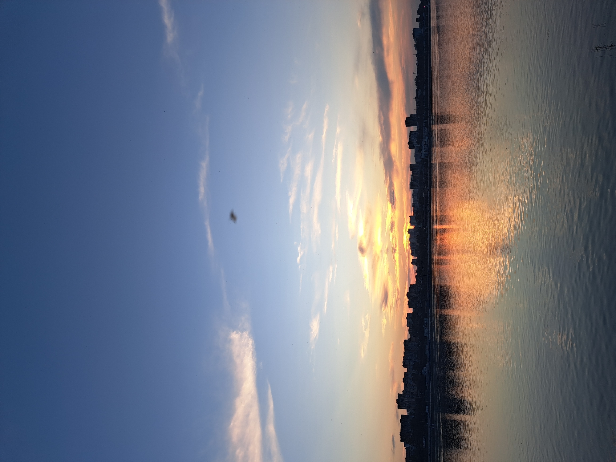 This image depicts a serene sunset over the Angara River in Irkutsk, Russia. A city skyline with numerous buildings, including apartment blocks, is visible in silhouette on the far bank. The sky is a gradient of deep blue at the top, transitioning to lighter blues and pinks and oranges near the horizon, illuminated by the setting sun. Wispy clouds are scattered across the sky, catching the warm light. The river's surface is calm, reflecting the colorful sky and the city lights, creating a mirror image of the scene above. The overall atmosphere is tranquil and picturesque, capturing a typical evening view of the city by the water.