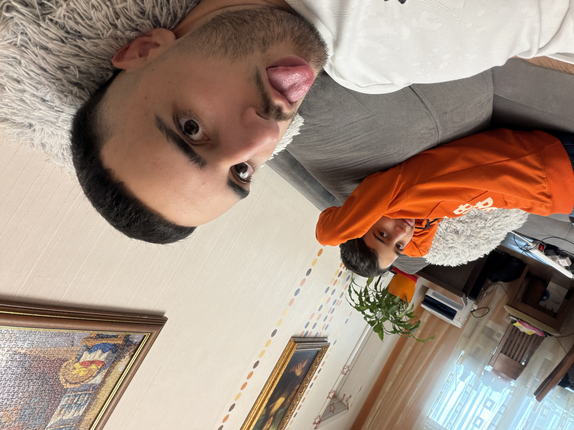 The image depicts two people, one adult and one child, in an indoor setting. The adult male, positioned in the upper-left, is lying on a gray sofa with a white shirt. He has dark hair and a beard, and his tongue is sticking out. A light-colored, fluffy pillow is behind his head. The child, wearing an orange sweatshirt, is lying on another fluffy pillow at the right part of the image.
The setting appears to be a home interior. In the background, there is a patterned wall with two framed pictures, one depicting a puzzle and the other showing a landscape. A potted plant and a window with sheer curtains are also visible in the background.
No information in the image indicates the location is in Plovdiv, Bulgaria. The description is purely based on the visual elements.