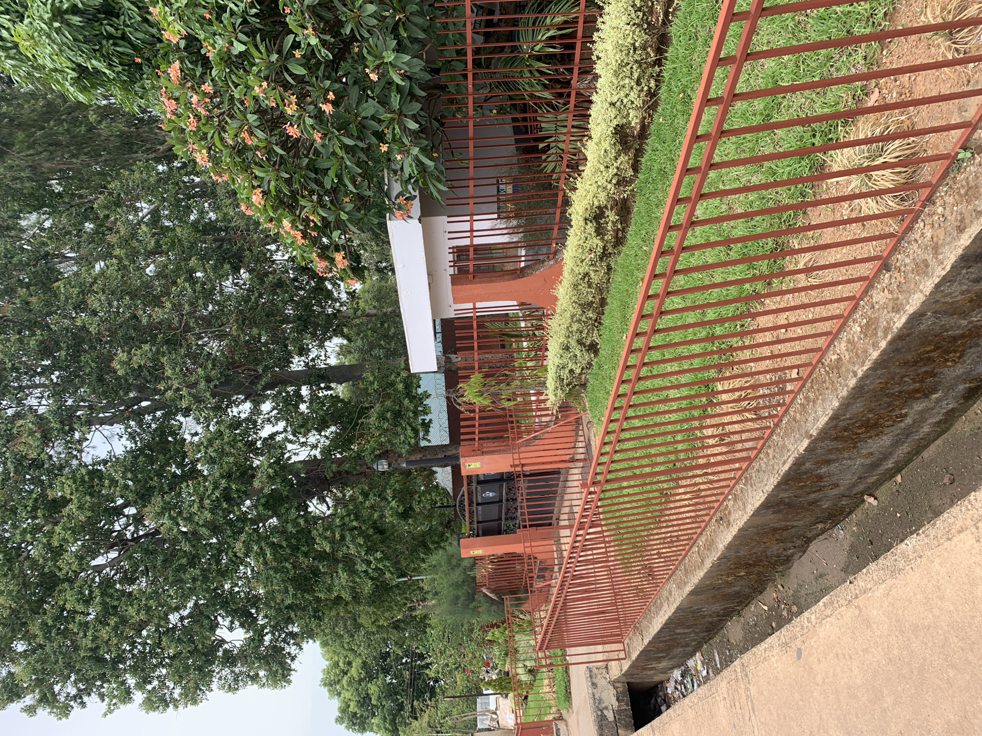 The scene depicts a residential or commercial building in Barakin Lerawi, Nigeria, partially obscured by trees and foliage. The building is behind a reddish-brown metal fence. There is a concrete culvert with garbage and water along the sidewalk. The image is taken at a low angle, with the camera pointed upwards toward the building. There are no people visible in the frame.