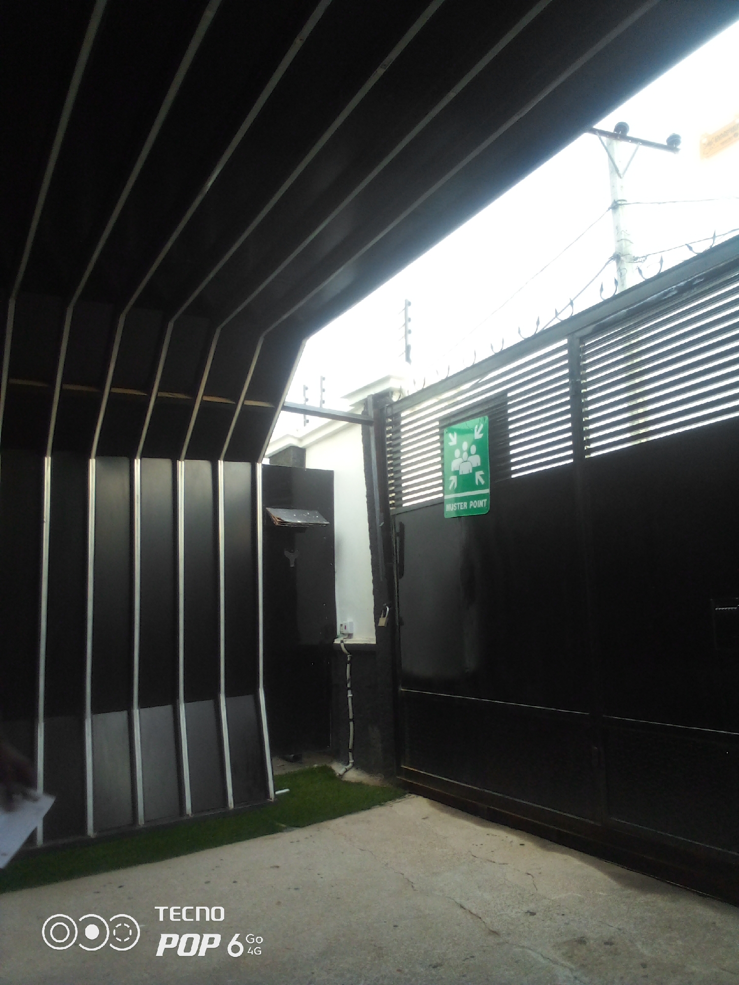 The image shows a black metal gate with horizontal slats in Howayi, Nigeria. A green sign with white icons and text reading "MUSTER POINT" is attached to the right gate panel. The gate is partially open, revealing a white building and what appears to be an electric fence on top of a grey wall. To the left of the gate, a section of a dark, ribbed structure is visible, possibly an overhang or part of the building. A small patch of green grass is located in front of the gate on the left. The ground in front of the gate is a concrete surface with some visible cracks. A mobile phone logo and text "TECNO POP 6 4G" are present in the bottom left corner of the image. The sky in the background is bright and overcast.
